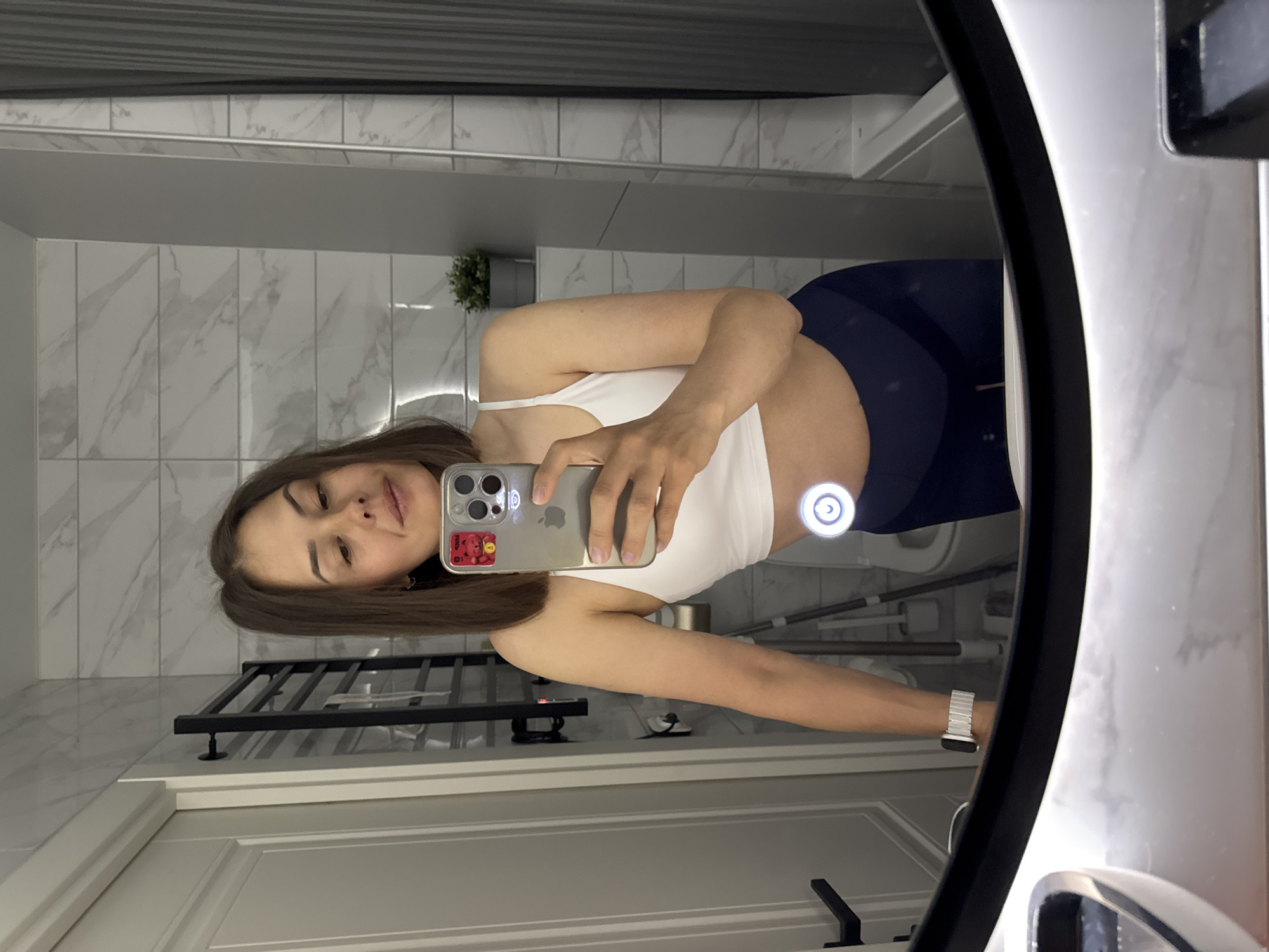 A woman is taking a selfie in a bathroom. The woman has brown hair and is wearing a white top and dark blue leggings. She is holding a silver iPhone in her right hand. Her left hand is visible, wearing a silver Apple watch. The bathroom has white marble-patterned tiles on the wall and a white door with a black handle. A black towel rack is mounted on the wall near the door. A small green plant sits on a shelf. The selfie is being taken in a round mirror with a white frame. The location is potentially in Chaikovsky, Russia.