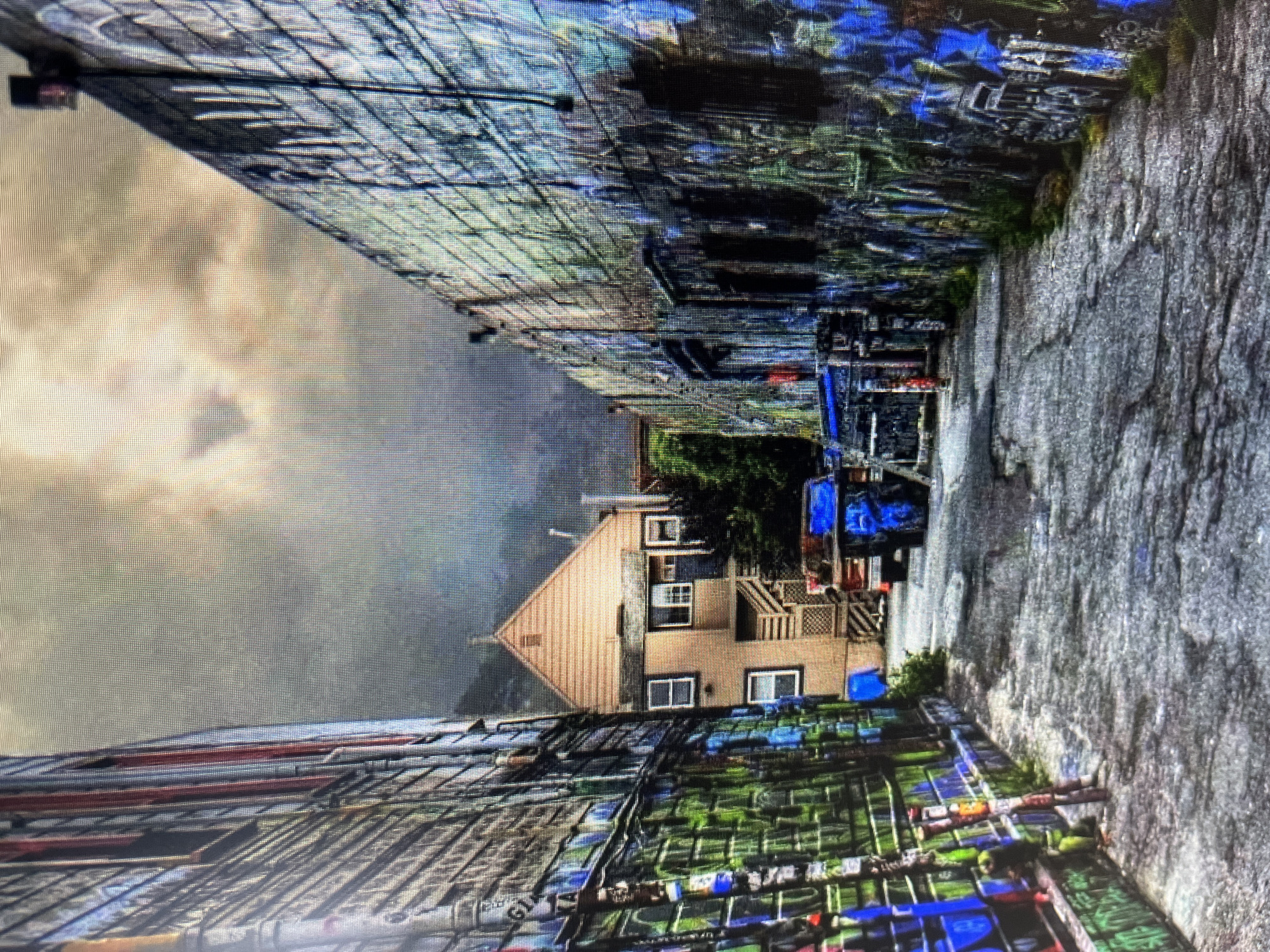 The image depicts a narrow, graffiti-covered alleyway. The walls on either side are densely covered in vibrant and chaotic spray paint, with blues, greens, and reds dominating the palette. The ground is a rough, textured surface, possibly asphalt or stone, and appears wet, suggesting recent rain or a damp environment.
In the background, a tan-colored house with multiple windows and a wooden exterior is visible, nestled between the alley walls. Stairs lead up to a deck or balcony. A blue structure, possibly a vehicle or equipment, is also present in the alley. The sky above is overcast and hazy, indicating a cloudy or possibly foggy day. The overall atmosphere is urban and somewhat gritty, with the bright graffiti providing a stark contrast to the muted tones of the sky and the house.
The perspective of the image is from within the alley, looking towards the house. The details of the graffiti are extensive, with various tags and designs layered on top of each other. There are some green plants growing in patches along the walls and near the house. The scene appears to be a back alley or a service entrance. There is no discernible text visible in the image. The time of day is unclear due to the overcast conditions.