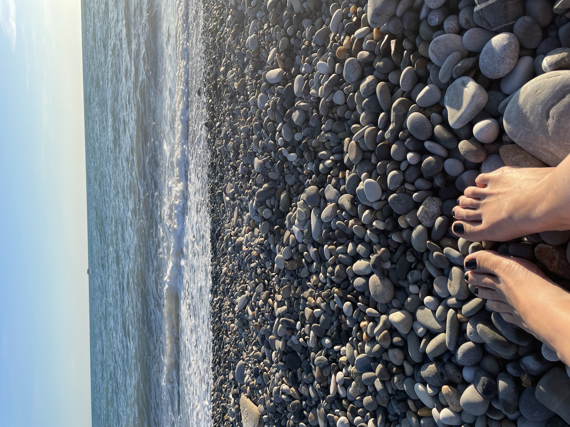 The image shows a close-up view of a pebbly beach with the ocean in the background. Two bare feet, with dark nail polish on the toenails, are in the foreground, resting on the pebbles. The ocean is calm with gentle waves breaking on the shore, and the sky is clear and blue with a hint of sunlight. A distant boat is visible on the horizon. The pebbles are of various sizes and colors, predominantly gray and brown. The scene suggests a peaceful moment at the beach.