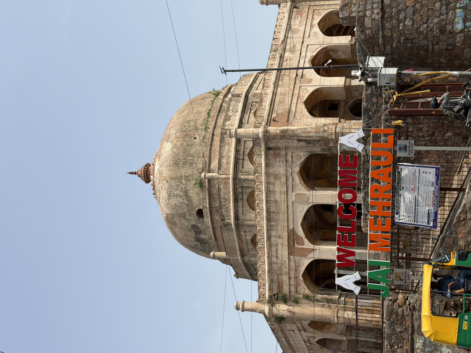 In New Delhi, India, the image captures a close-up view of the tomb of Adham Khan, a significant historical monument in Mehrauli. The sun-drenched day showcases the weathered stone structure, featuring a prominent dome and arched colonnades, with hints of greenery growing on its upper sections. In the foreground, a vibrant "WELCOME JAI MEHRAULI" sign is prominently displayed, with the letters in red and green, set against the backdrop of the historical building. Below this, a yellow and green auto-rickshaw is parked, with a driver seated inside. A person is seen on a motorbike behind a metal gate, wearing a backpack and a helmet. Several utility poles and wires are visible, indicating the presence of modern infrastructure in the vicinity of this ancient monument.