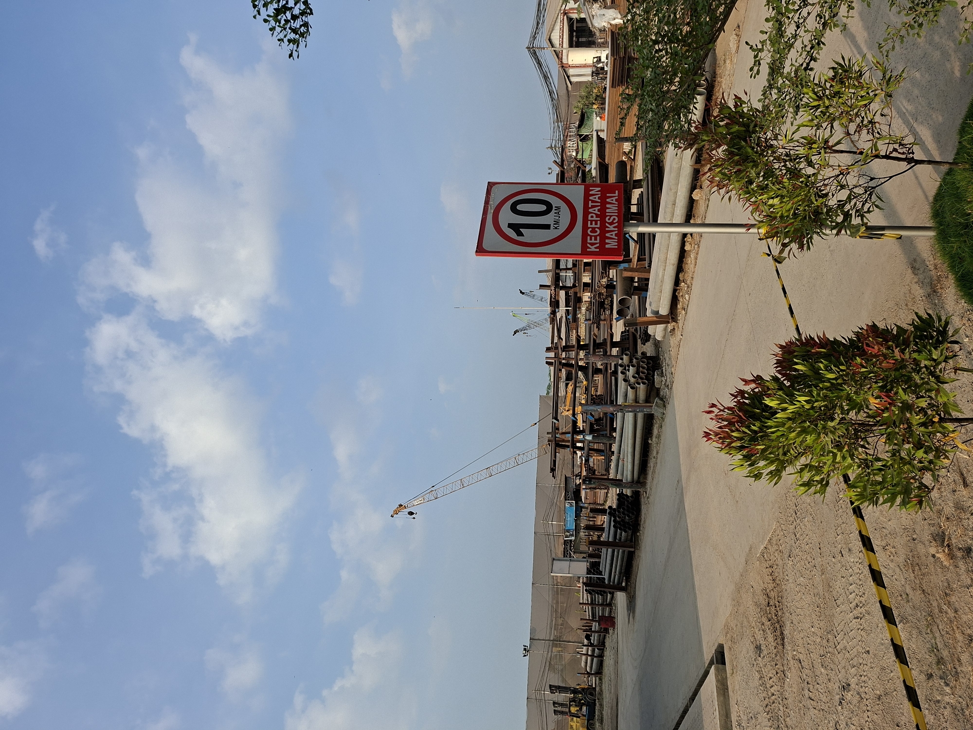 The image depicts a construction site in Loa Buah, Indonesia. A bright red and white speed limit sign, indicating a maximum speed of 10 km/h, is prominently displayed in the foreground. The sign reads "KECEPATAN MAKSIMAL" below the numerical speed limit. Behind the sign, various construction materials, including long pipes and metal bars, are stacked neatly. In the background, a large construction crane is visible against a bright blue sky with scattered white clouds. The ground is a mix of dirt and a paved surface. There are no people or vehicles clearly visible in the image. The overall scene suggests an active but regulated work environment.