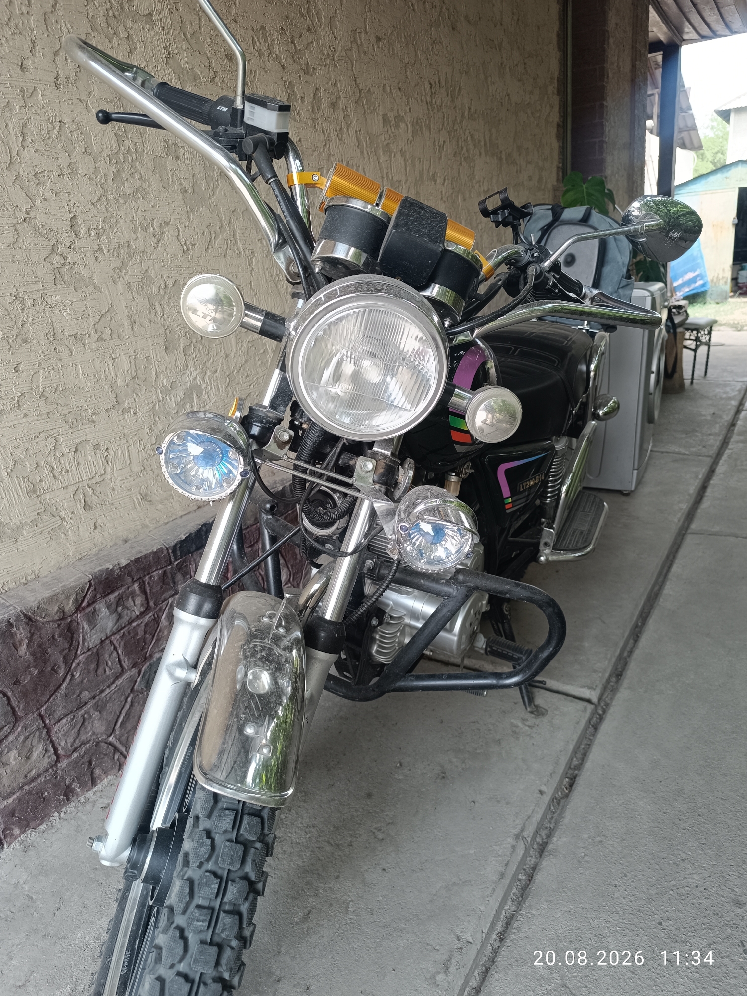 The image shows a black motorcycle parked outdoors, leaning against a textured wall. The motorcycle has a large circular headlight, two smaller auxiliary lights on either side of the main headlight, and orange turn signals. It features chrome handlebars, mirrors, and various other chrome accents. The motorcycle is equipped with a black seat, footpegs, and a protective engine guard. The ground beneath the motorcycle is concrete, with a visible expansion joint. In the background, parts of a wooden overhang and a building with a light-colored wall are visible. The timestamp "20.08.2026 11:34" is present in the lower right corner. The scene appears to be located in Qaskelen, Kazakhstan, likely in a residential or commercial outdoor area, possibly a courtyard or garage entrance. There are no people visible in the image.