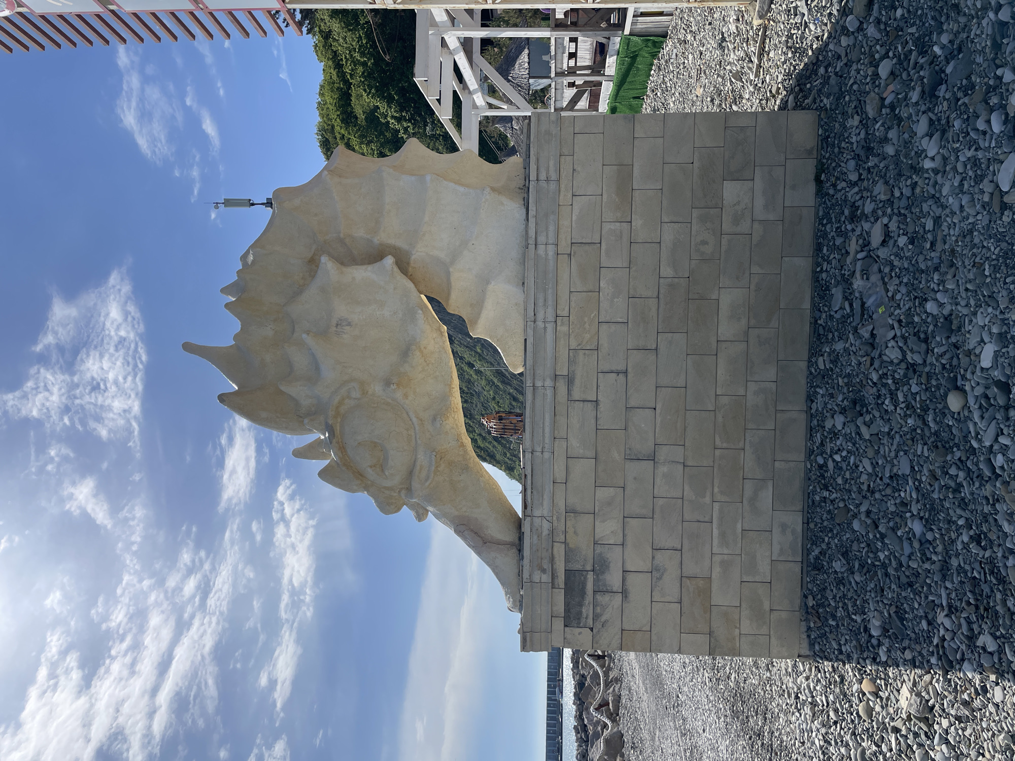 The image features a sculpture of a seahorse in Olginka, Russia. The seahorse is mounted on a stone-block base. The background includes a blue sky with clouds, foliage, a wooden pergola, and a building structure. The ground consists of pebbles and a tiled area.