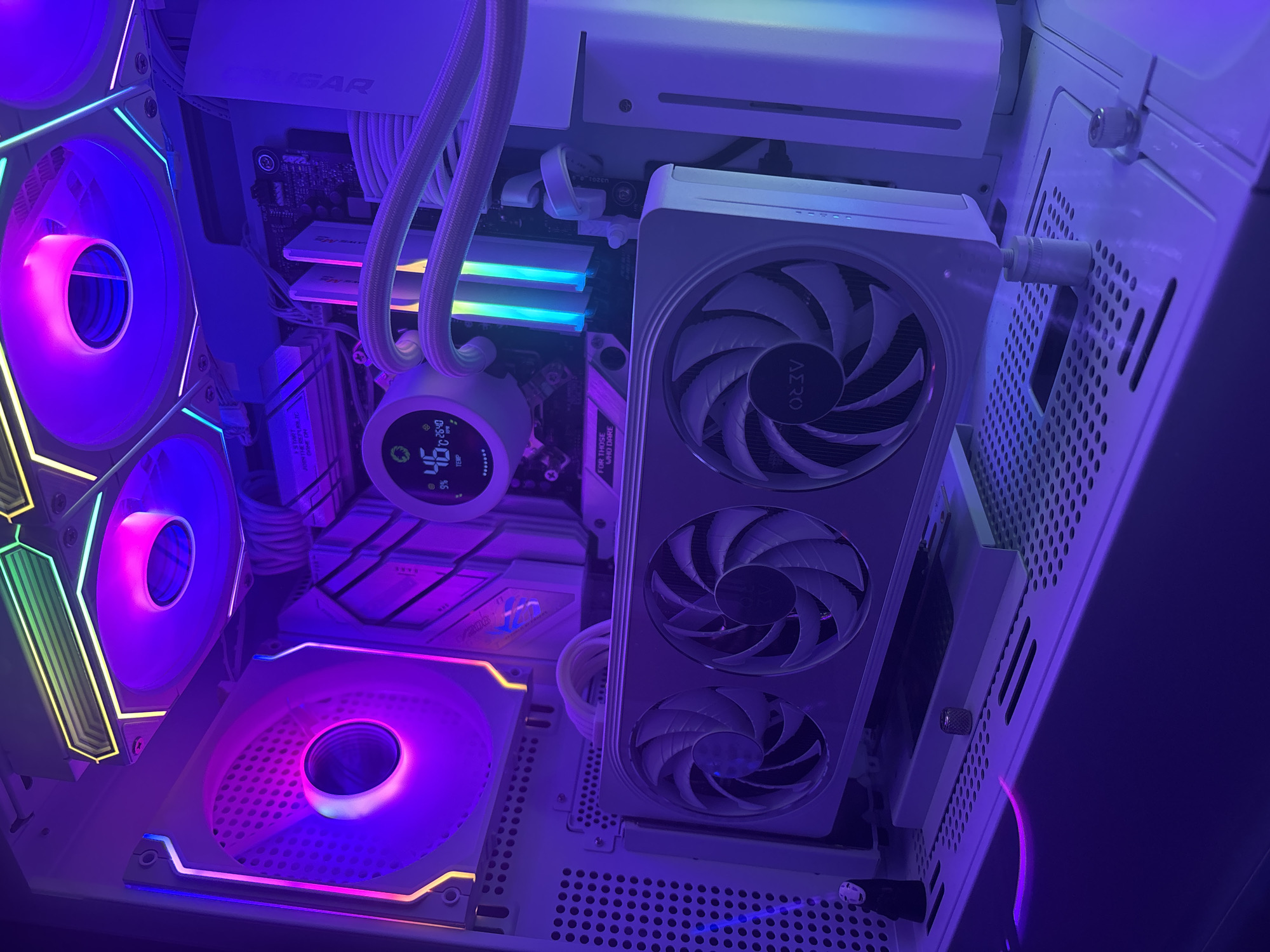This is a close-up, high-angle shot of the interior of a white computer case, bathed in purple and blue LED lighting. The components are meticulously arranged and illuminated. In the foreground, a white graphics card with three large fans dominates the lower half of the image. The fans have a stylized, curved blade design and a black logo in the center. Above the graphics card, a circular LCD display shows system information, including temperature readings of 46°C and fan RPMs of 2540. White braided cables snake throughout the build, connecting various components. Two tall RAM modules with RGB lighting, displaying cyan and blue hues, are visible behind the graphics card. The top of the case features two large, illuminated fans with RGB lighting, casting purple and pink glows. The overall impression is a clean, modern, and visually striking computer setup. The location is unknown.