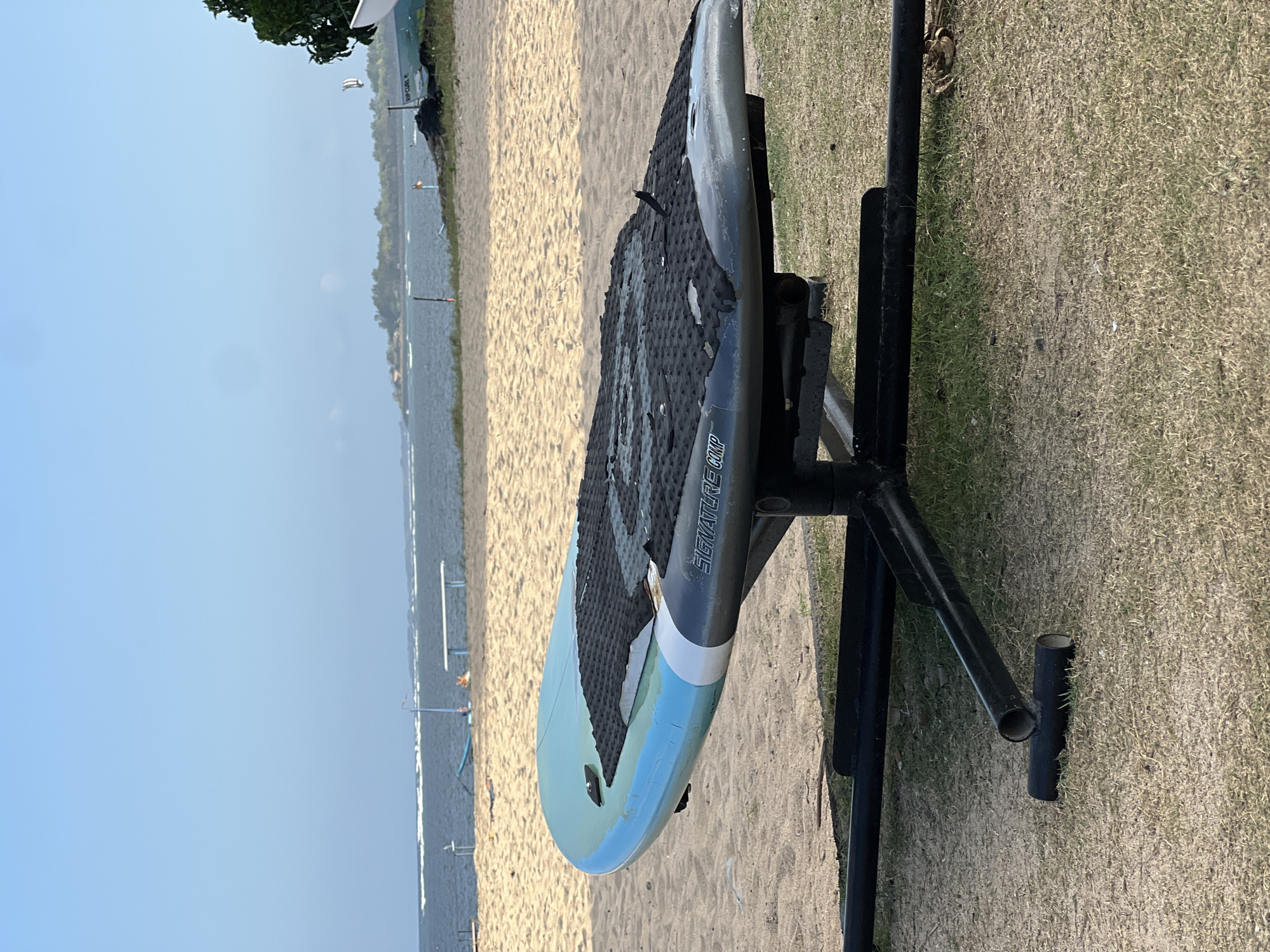 A surfboard rests on a black metal stand on a grassy area next to a sandy beach. The surfboard is light blue with black and white accents and has a black grip pad on top. The ocean is visible in the background with gentle waves and a clear sky. In the distance, there are some trees and buildings. The scene appears to be in Denpasar, Indonesia.