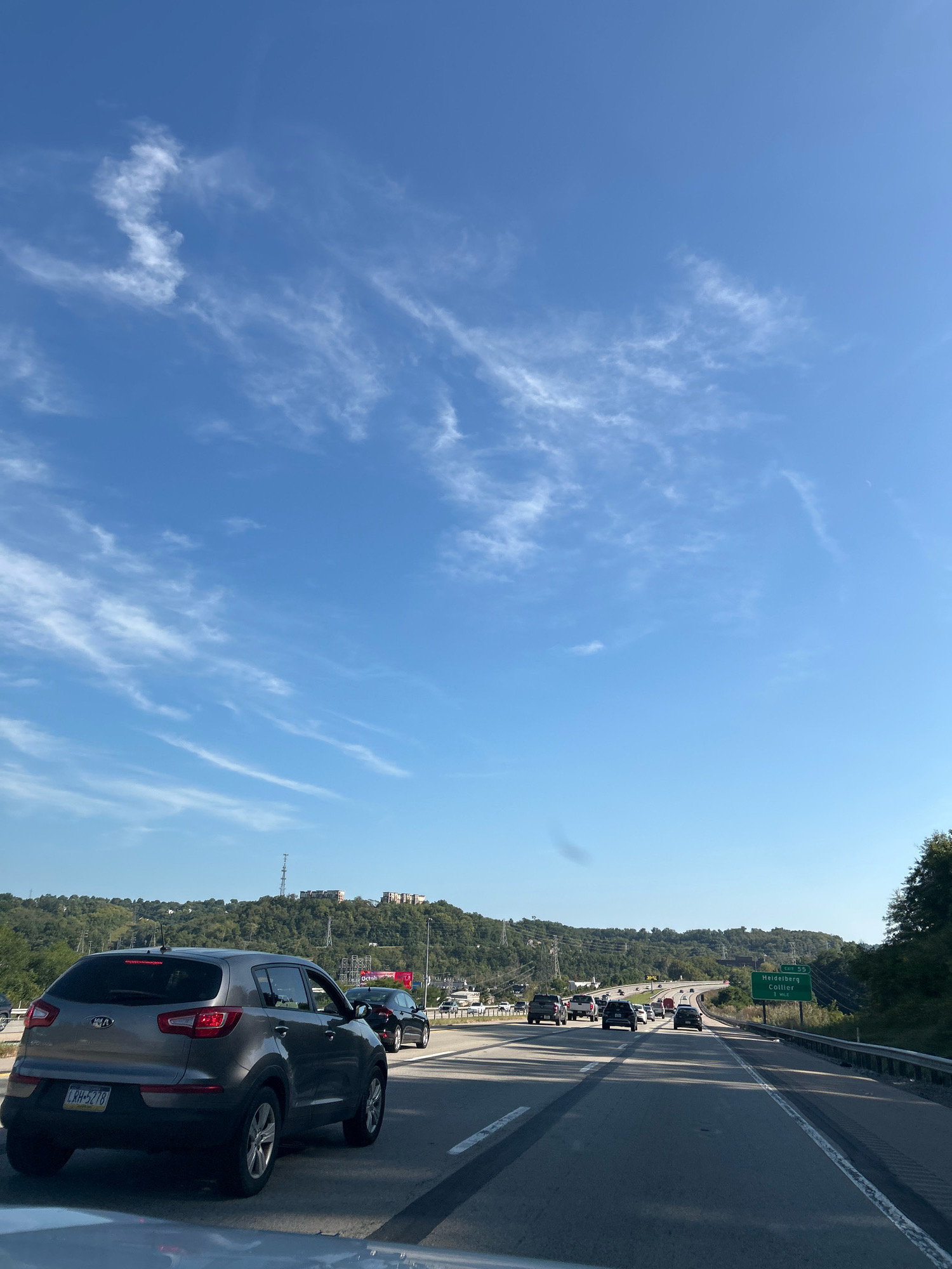 The image shows a highway scene in Collier Township, United States of America. The sky is blue with scattered white clouds. There is traffic on the highway, with several cars visible. In the foreground, a gray SUV is in the right lane. In the background, there are hills covered with trees. A green highway sign indicates "Exit 55 Heidelberg Collier 1 Mi". The scene appears to be during the daytime.