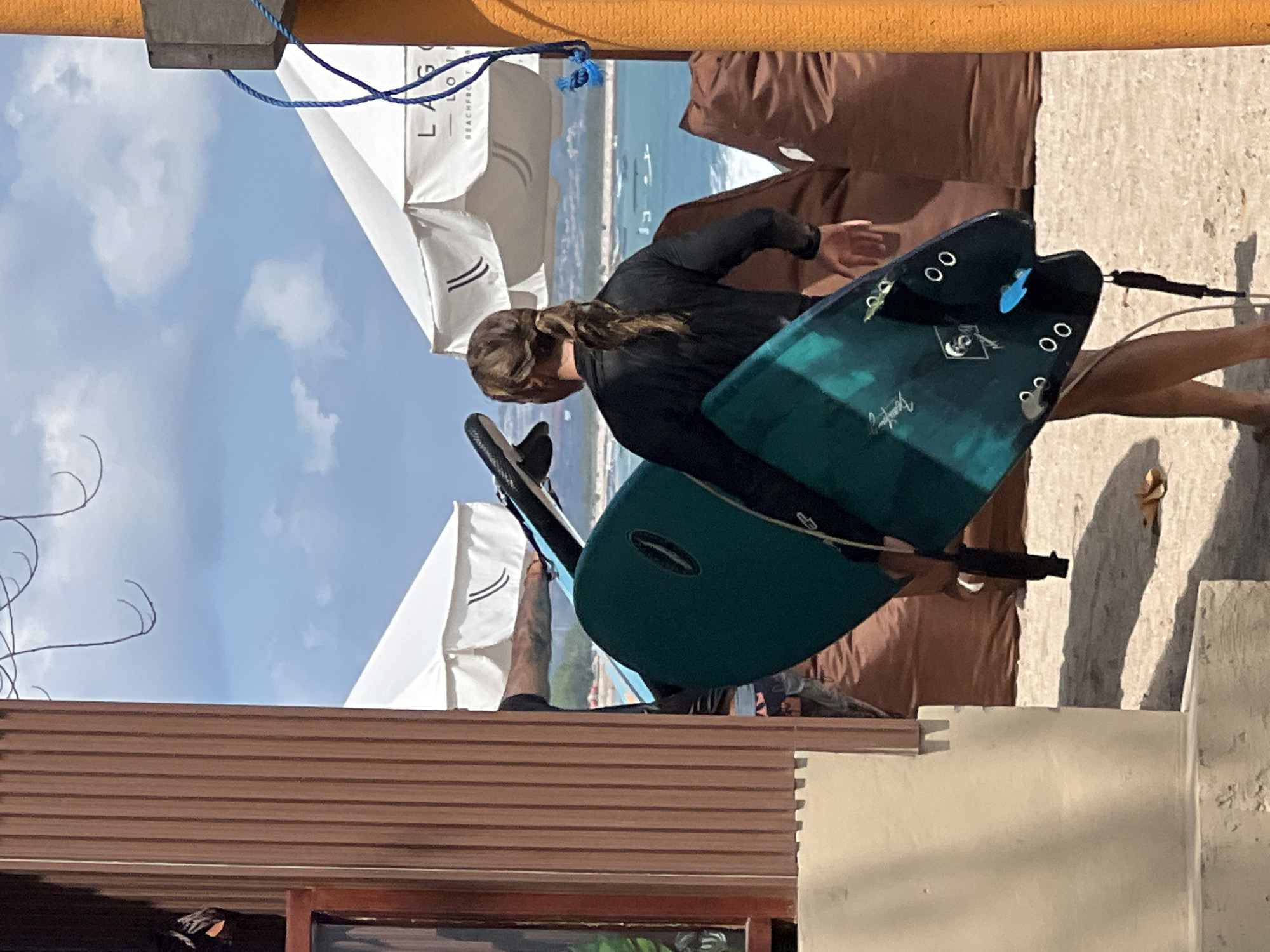 In Kuta, Indonesia, a person with long hair tied in a ponytail walks away from the camera on a sandy beach. They are wearing a black long-sleeved rashguard and are carrying two surfboards. The surfboards are teal and black with white lettering. In the background, two white umbrellas provide shade, and beyond them, the turquoise ocean stretches out under a blue sky with scattered clouds. Beach chairs are visible behind the umbrellas, and a brown wall, likely part of a beachside establishment, is to the left of the frame. An orange pole with a blue rope is on the right side of the image.