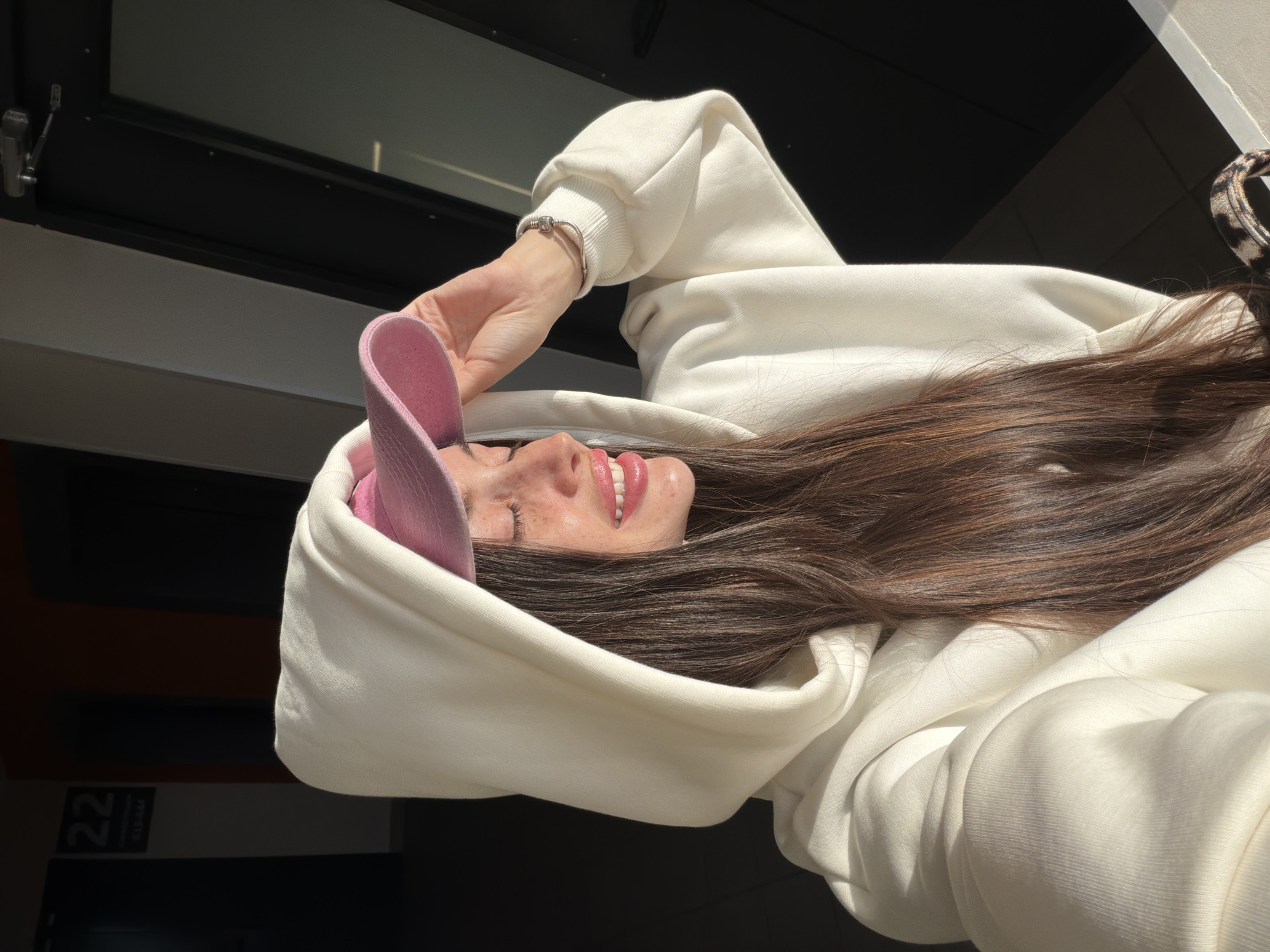 A young woman in a white hoodie and pink baseball cap, with long brown hair, is holding the brim of her cap. She is smiling. The background is partially obscured, showing a window and a building exterior with the number "22" visible. Based on architectural style, materials, and color palette, the location may be in the city of Saint Petersburg, Russia.