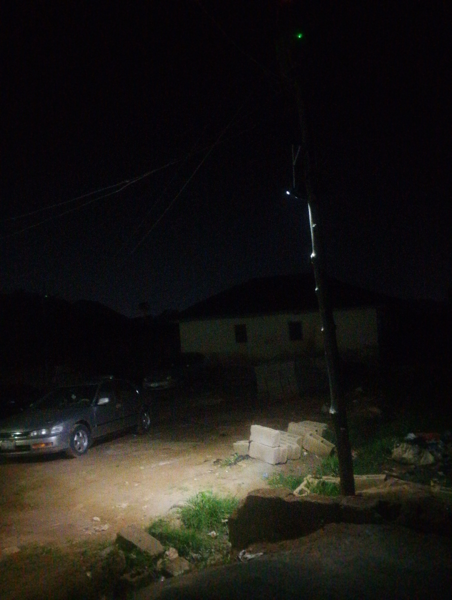 The image depicts a dimly lit outdoor scene at night, likely in the city of Jos, Nigeria. In the foreground, a sedan is parked on an unpaved surface, illuminated by artificial light. The car appears to be silver or gray. To the right of the car, a pile of concrete blocks is visible, along with some patchy grass and debris. In the background, a building with a dark roof and visible windows is partially obscured by darkness. A utility pole stands prominently in the center-right of the frame, with a small green light near its top. Overhead, thin wires are strung across the dark sky. The overall impression is one of a quiet, nocturnal urban or semi-urban environment.