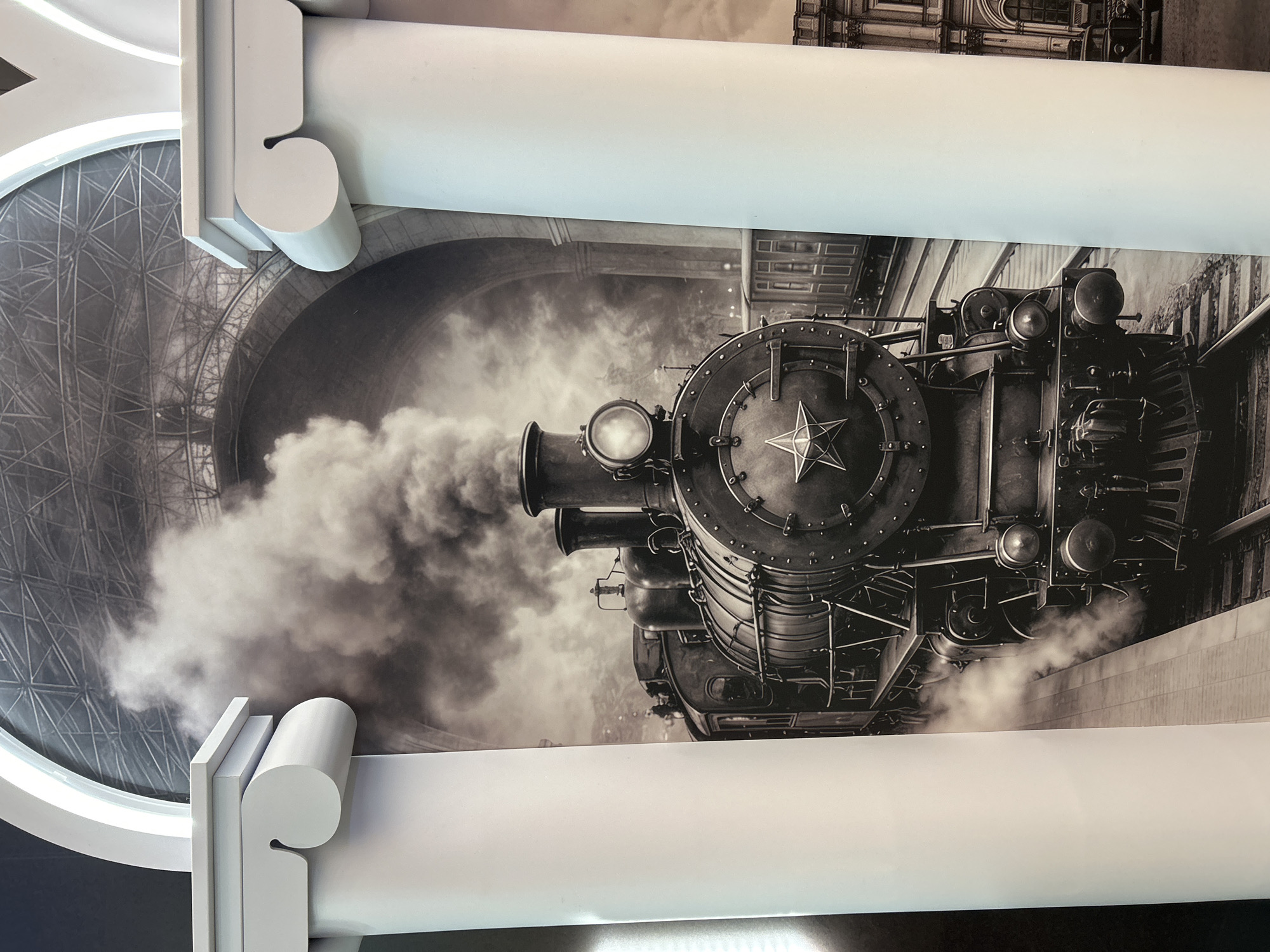 The image captures a dramatic, black-and-white depiction of a vintage steam locomotive, likely a mural or ceiling art, framed by ornate white architectural elements. The steam engine is the central subject, seen head-on with a slight angle to the right, heavily emitting thick plumes of smoke and steam from its smokestack and sides, suggesting movement or immense power. A prominent five-pointed star emblem is visible on the front of its smokebox door, above the large, illuminated headlight.
In the background of the artwork, architectural structures are visible: a large, arched opening with intricate lattice-work or scaffolding on the left, and old-style buildings or station platforms on the right. The overall aesthetic of the train image evokes a historical, industrial era.
The entire black-and-white artwork is surrounded and partially obscured by prominent, bright white architectural features. These include thick, stylized columns or beams with both rounded and angular decorative details, which appear to be part of the actual building's interior structure, possibly framing a large ceiling installation or high wall. The bright white of these elements strongly contrasts with the dark tones of the steam train. No visible text is present in the image.
