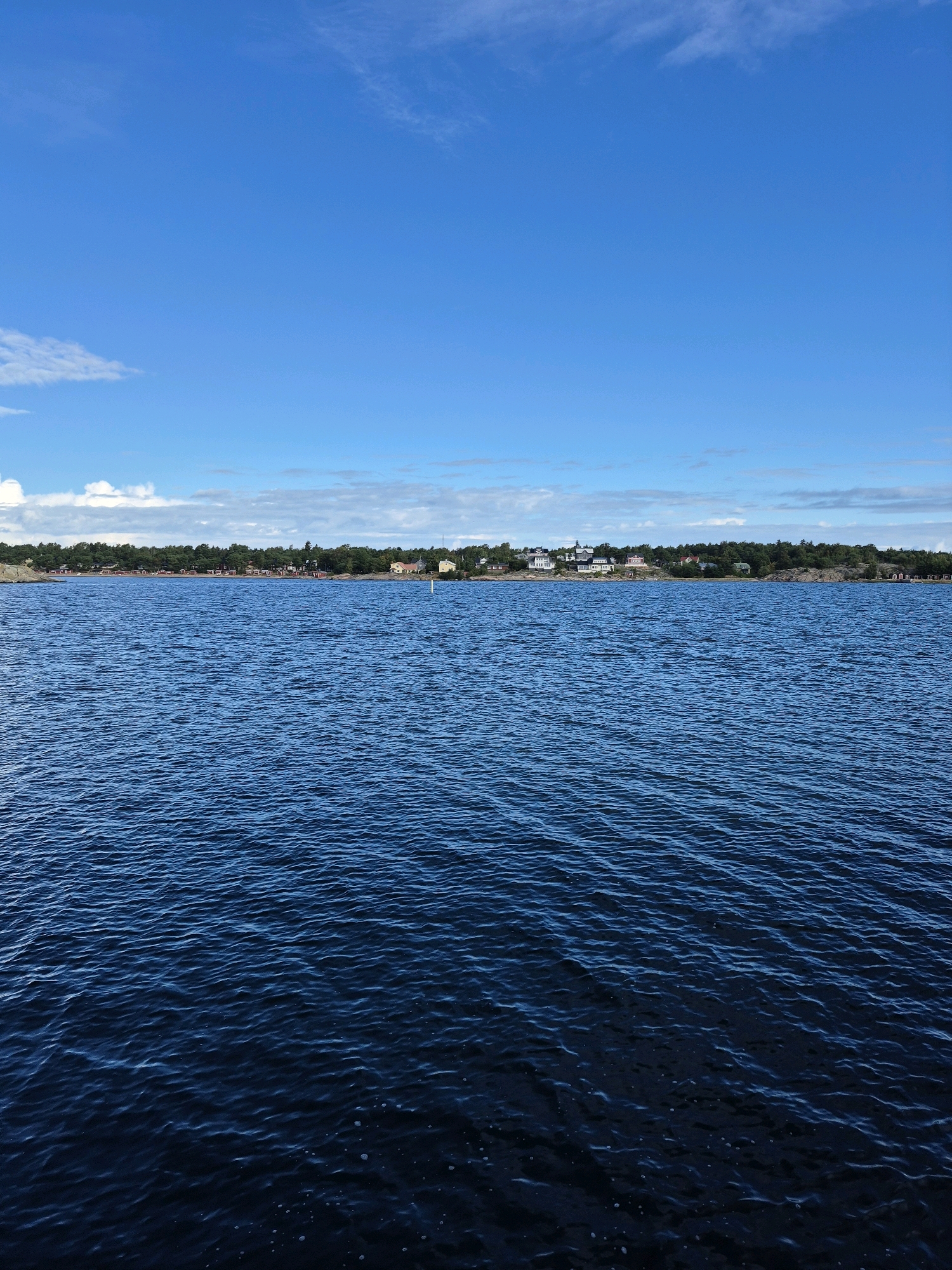 The image depicts a serene seascape with a clear blue sky overhead. The foreground is dominated by a vast expanse of deep blue water, with gentle ripples indicating a calm surface. In the mid-ground, a line of rocky land stretches across the horizon, densely covered with dark green trees. Beyond the trees, a small cluster of buildings, likely houses and other structures, are visible, suggesting a coastal settlement. A white buoy stands in the water near the shore. The location is identified as being in Hanko, Finland. The scene is devoid of any people or visible human activity, emphasizing the tranquility of the natural environment and the settlement. The lighting suggests it is daytime, likely late morning or early afternoon, with the sun high in the sky, contributing to the bright blue of both the water and the sky.