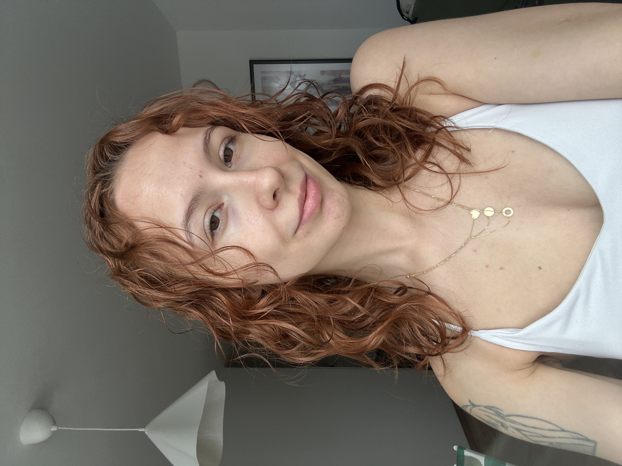 A young woman with red curly hair is looking at the camera. She is wearing a white tank top and a gold necklace with small circular and heart-shaped charms. She is in a room with gray walls. A white lampshade is visible in the lower left corner. A framed picture hangs on the wall behind her. A tattoo is visible on her upper arm. The image was not taken in Novi Sad, Serbia.