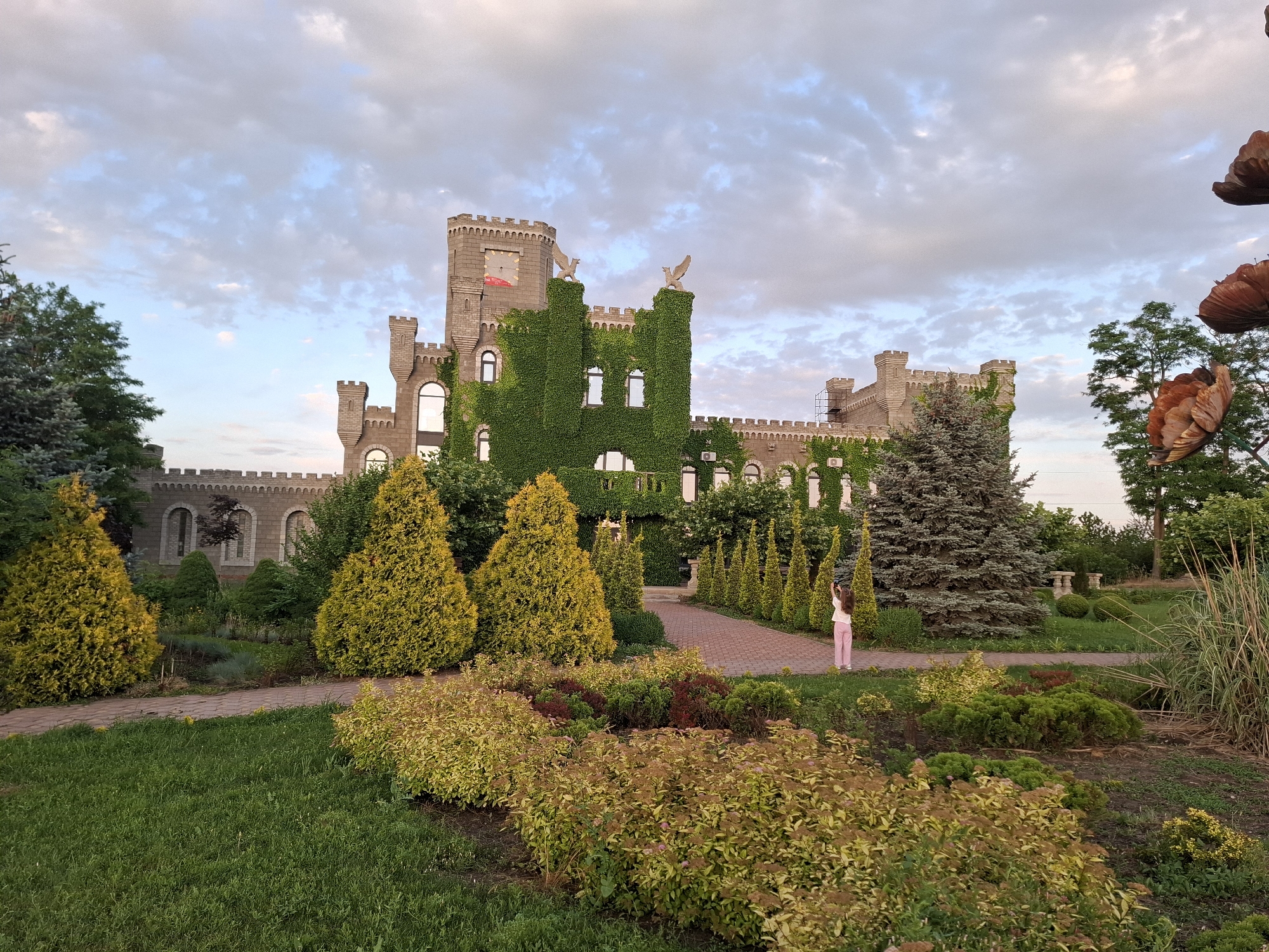 A large, stone castle-like building, partially covered in ivy, is the main subject of the image. The building features multiple towers with crenellated tops, arched windows, and some decorative eagle statues on its upper sections. In front of the building, a paved walkway leads through a manicured garden with various types of trees and shrubs, including vibrant yellow conifers and colorful flowering bushes. A young person, wearing pink pants and a white top, stands on the walkway near the right side of the image, holding a phone up as if taking a picture. The sky above is partly cloudy with hints of blue. The overall scene suggests a park or tourist attraction. The location is identified as Korenovsk, Russia, implying this is likely a landmark or park within that city.