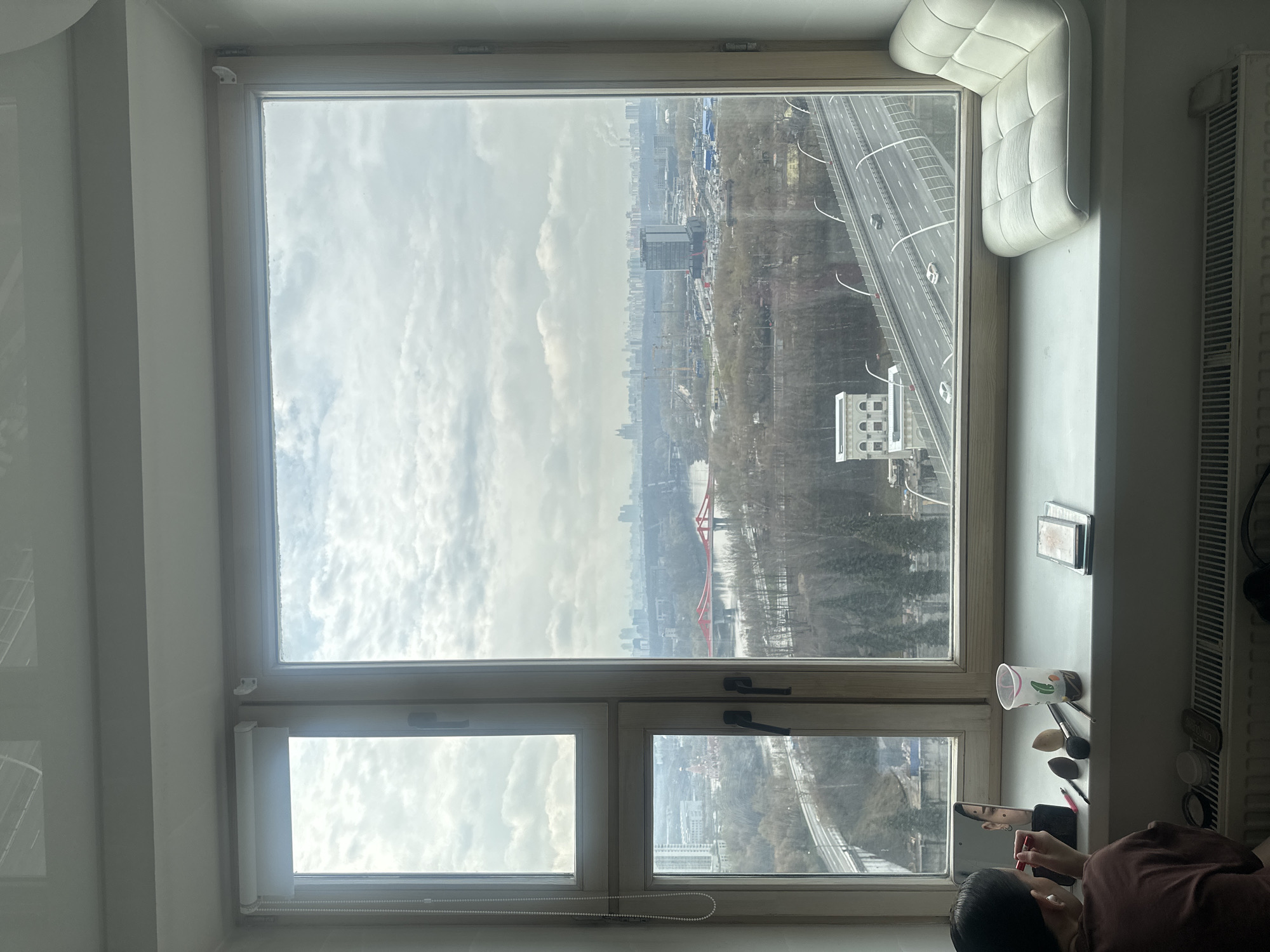 An interior view through a large window, showing a cityscape in the background, likely in Khimki, Russia. The view includes a multi-lane highway with moving vehicles, and a red bridge in the distance. Visible buildings are interspersed with patches of trees. The sky is overcast.
On the windowsill, various items are scattered, including makeup brushes, a cup, and cosmetic containers. A person is partially visible in the lower right corner, appearing to be applying lipstick while looking at a handheld device. A cushioned window seat is situated in the upper right corner. A radiator is present on the right side of the frame. The window is divided into multiple panes.