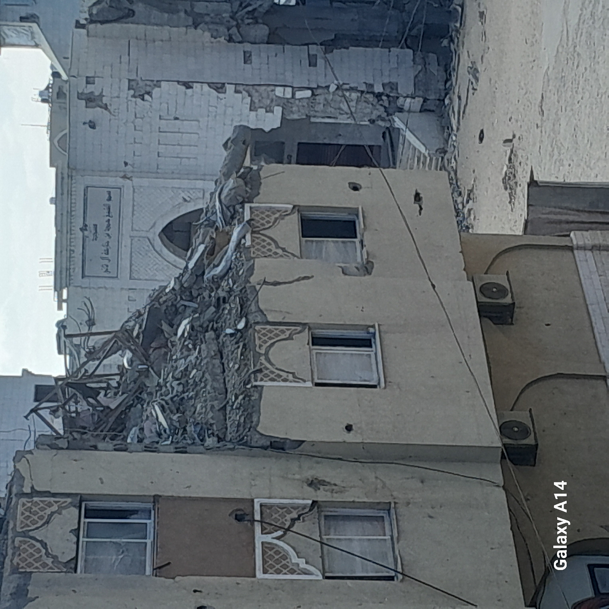 The image shows a partially destroyed building in Khan Yunis, Palestinian Territories. The building appears to be a multi-story structure made of concrete and brick, with visible damage from what appears to be an explosion or shelling. Debris, including concrete chunks and twisted metal, is piled on top of the building and surrounding area. Windows are broken, and sections of the walls are missing. The architecture suggests a residential or commercial building, with some decorative elements above the windows. The ground around the building is sandy and covered with rubble. A sign with Arabic text is visible on an adjacent structure. There are no people or animals visible in the image. The overall scene depicts significant structural damage and destruction.