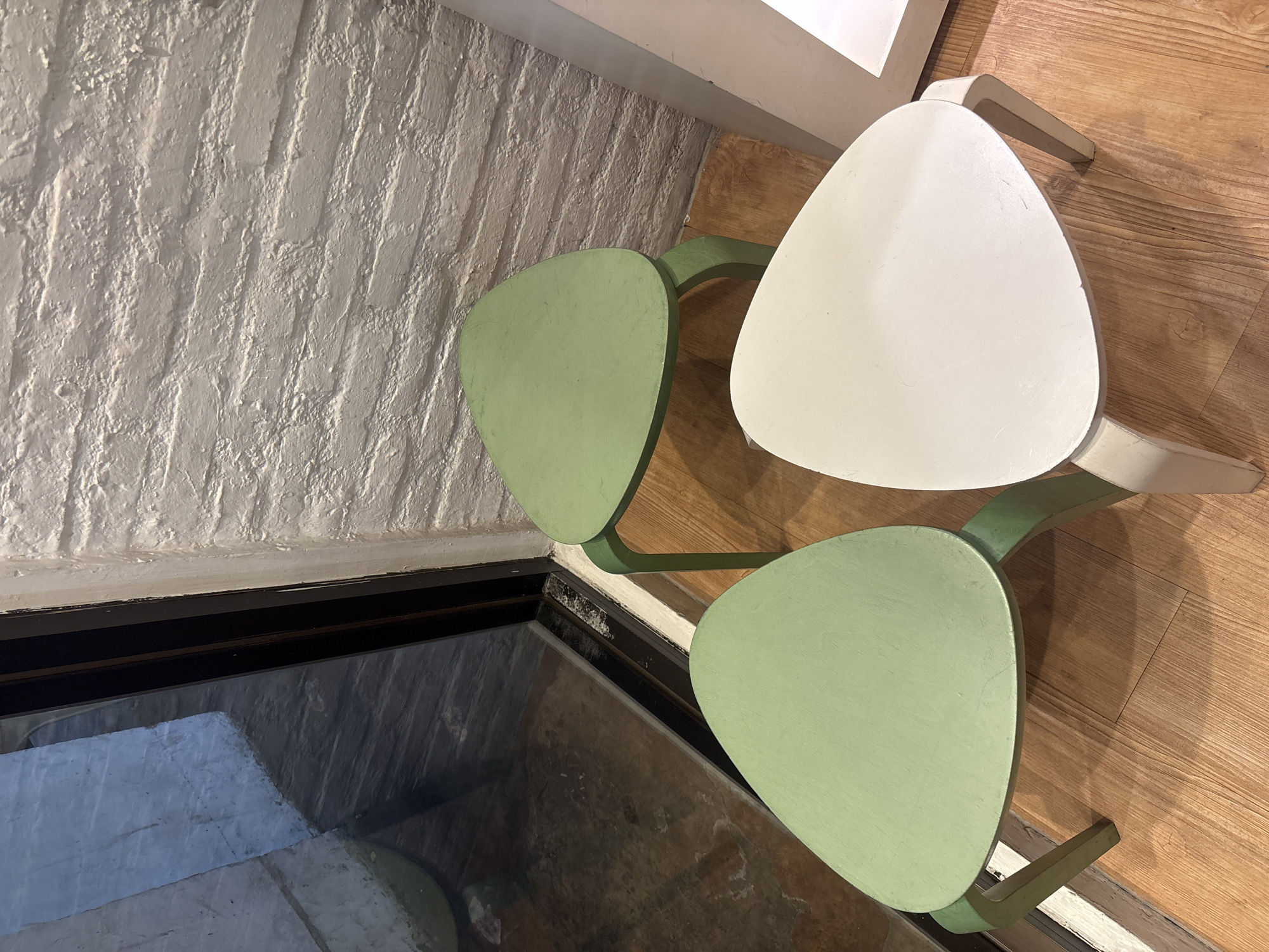 The image shows an overhead view of two chairs with triangular seats positioned near a window and a brick wall. One chair has a white seat, while the other two have light green seats. The chairs are likely indoors on a wooden floor. The location is unknown and cannot be determined from the image.