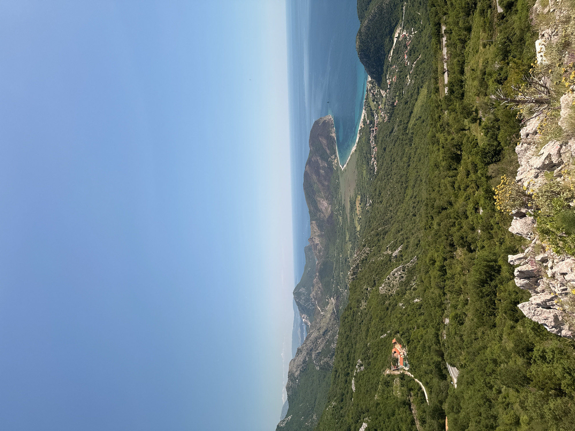 An aerial view from the mountains in Novoselje, Montenegro, shows the blue Adriatic Sea, a mountainous peninsula, lush green vegetation and a few structures among the trees. The sky is a gradient of light to dark blue. The terrain slopes downward from the right to the left. Exposed grey and white rock formations are visible on the upper right side. Below those, dense green trees extend down the slope, with a reddish-roofed building and winding road visible. The peninsula extends into the sea, a sandy beach stretches along its base.