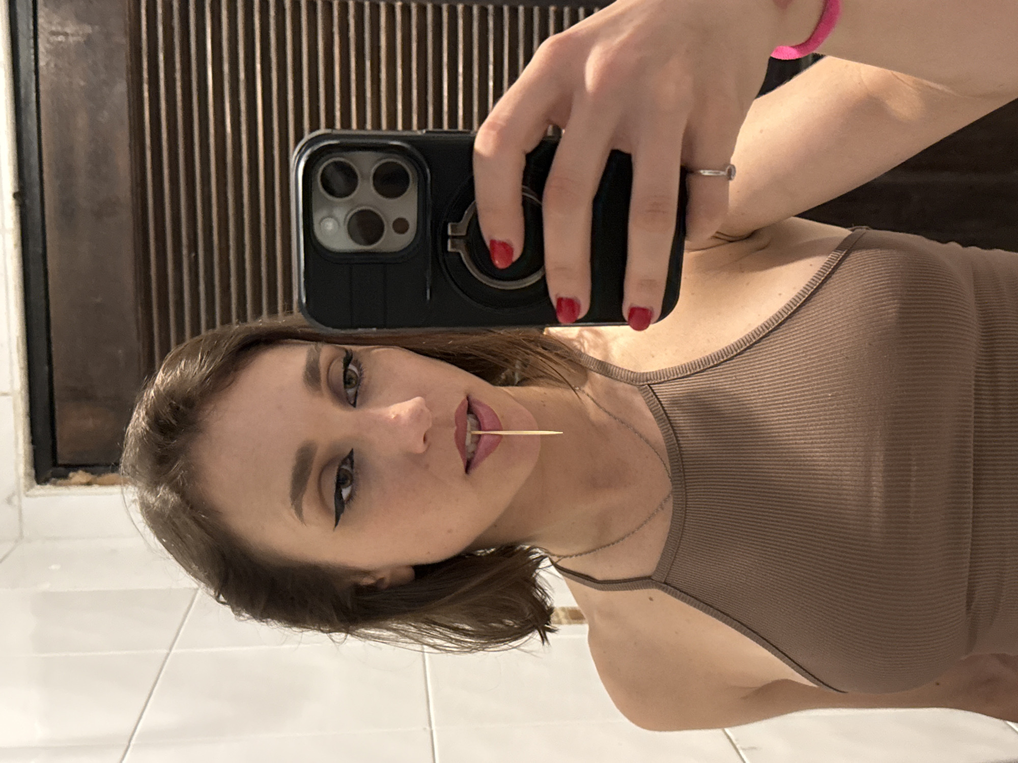 A woman is taking a mirror selfie in a bathroom. She has brown hair and is wearing a brown ribbed tank top. She has on winged eyeliner and a gold toothpick is placed between her lips. She is holding up a black smartphone in her right hand, which has a ring on the back. She has red nail polish on her fingernails. A pink bracelet is visible on her left wrist. The background shows white tiled walls and a wooden door with horizontal slats.