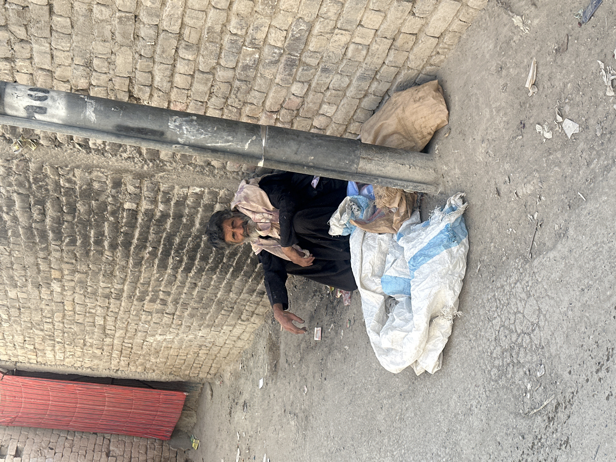 A man is seen lying on the ground next to a brick wall and under a metal pipe. He is dressed in dark clothing and has a light-colored cloth draped around his neck and shoulders. He is lying next to a white bag and a brown bag. His arm is extended. The ground is a light gray color with debris scattered on it. A red roll is visible on the left side of the image. This location is in Quetta City Tehsil, Pakistan.