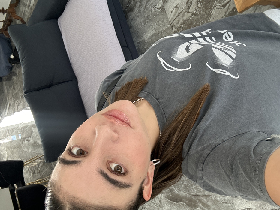 A woman is pictured from a high angle, wearing a gray graphic tee. She has brown hair and brown eyes, with a silver hoop earring visible. Behind her is a gray sofa with a dark gray and textured white pillow. The background also includes a tiled floor, a glimpse of a window, and part of a black chair.