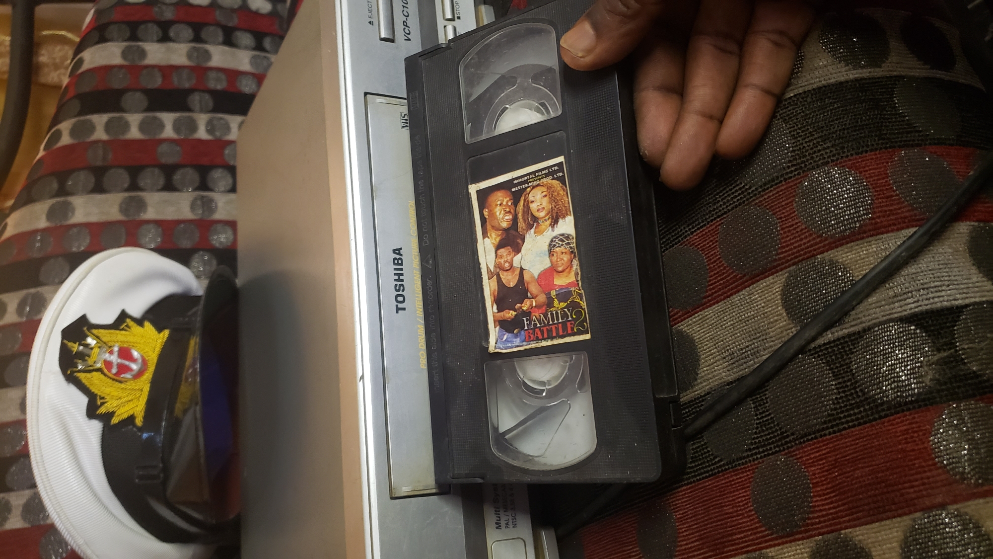 The image shows a VHS tape labeled "FAMILY BATTLE 2" being held by a person's hand. The tape has a sticker on it with the movie's title and images of the actors. In the background, a Toshiba VCP-C100 VHS player is visible, along with a naval officer's hat. The scene appears to be indoors, possibly in a living room, with a patterned couch in the foreground. The lighting suggests it is daytime. The location is not specified in the image itself, nor is there any visual information to place it in Narayi, Nigeria.