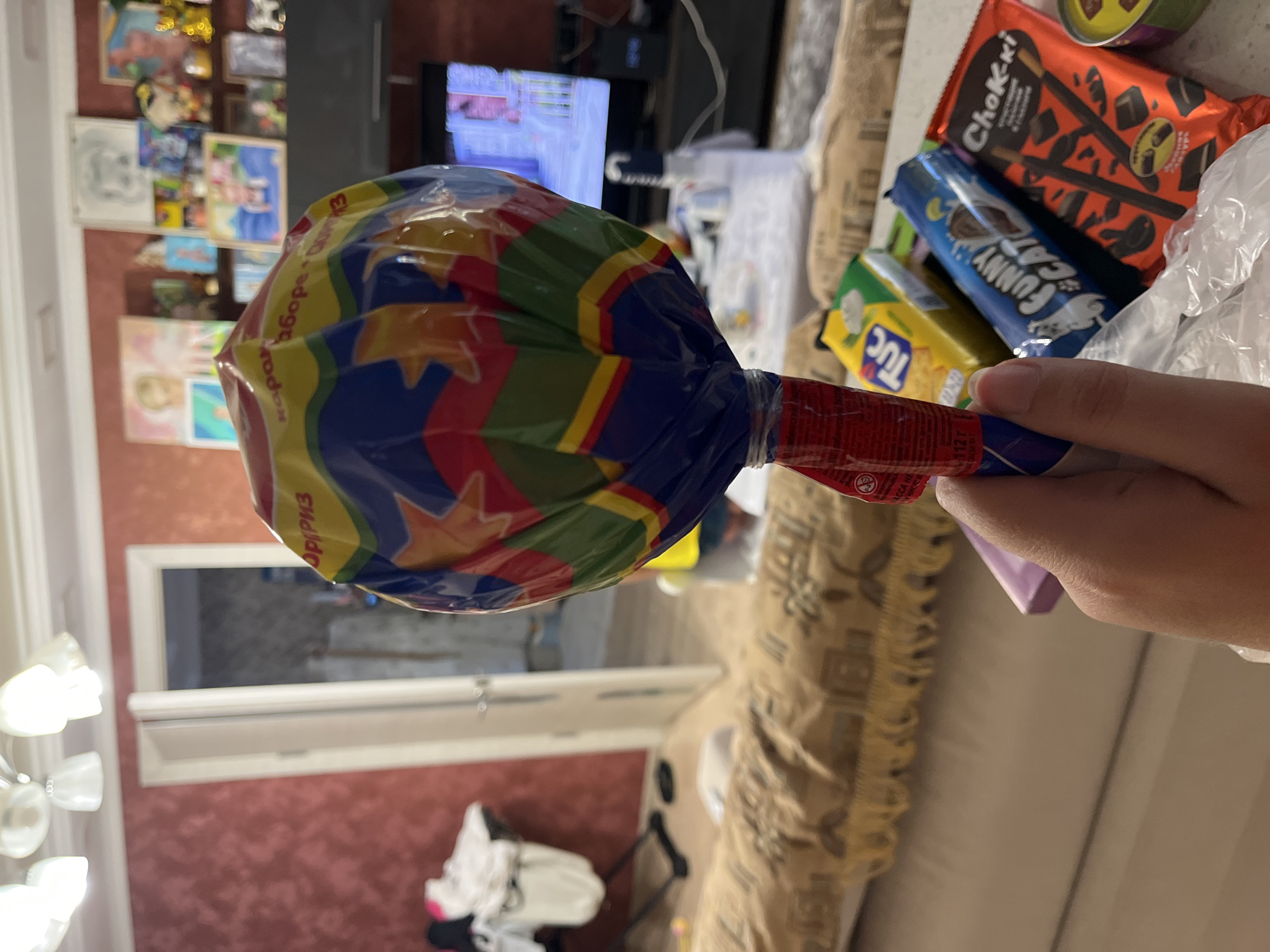 A hand holds a large, brightly colored lollipop wrapped in cellophane. The lollipop's wrapper features a pattern of stars and zig-zag lines in shades of blue, yellow, red, and green. A red label is attached to the stick of the lollipop. In the background, a living room environment is visible. A couch with a patterned throw blanket is in the foreground. Beyond the couch, there is a doorway leading to another room, a television set, and shelves filled with various items including framed pictures and decorative objects. Several snack packages are arranged on a counter or table in front of the couch, including a "Tuc" cracker package, a "Funny Cat" snack, and a "Chok-ki" chocolate stick package. The lighting suggests an indoor setting during the day. The presence of Russian text on some of the snack packaging indicates the location is likely in Russia.