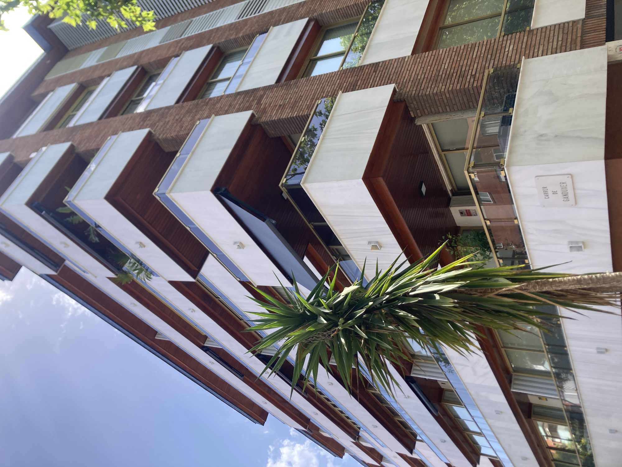 This media file shows a low-angle view of a modern building in Barcelona, Spain, with a bright blue sky and scattered clouds overhead, suggesting fair weather. The building features a distinctive facade with alternating bands of textured brickwork and smooth, light-colored panels that appear to be marble. Long, narrow windows are interspersed across the levels, reflecting the surrounding environment, including trees and sky.
Several cantilevered balconies or architectural extensions jut out from the building, creating a layered effect. These extensions have white or light-colored soffits and are framed by darker wooden elements, with reflective glass panels that mirror the sky and greenery. A large, spiky green plant, possibly a yucca or palm, is prominently positioned in the foreground, its fronds reaching upwards towards the building and sky, adding a natural element to the urban architecture.
One section of the building has a white plaque with dark text that reads "CARRER DE GANDOUKER". This is a clear location cue, indicating the name of a street. The overall impression is one of contemporary architecture, with a blend of natural and man-made materials, set against a clear daytime sky. There are no people visible in the image.