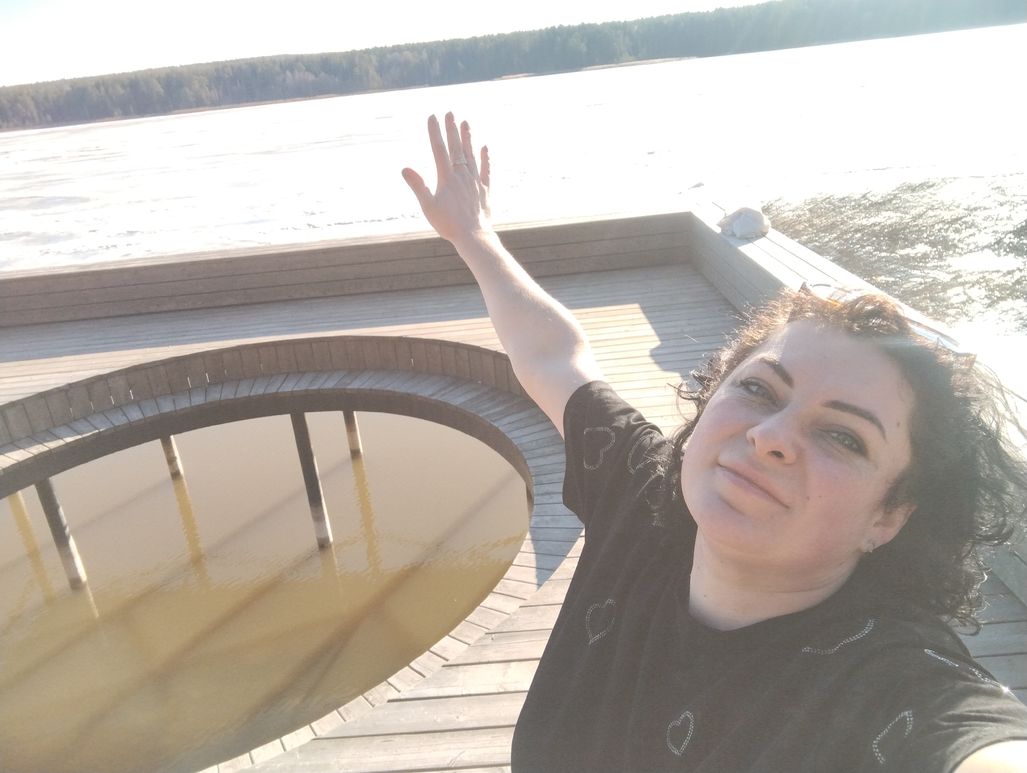 A woman takes a selfie outdoors, on a wooden deck overlooking a body of water. The deck features a circular opening into the water below. The woman is wearing a black t-shirt decorated with heart patterns. Her right arm is raised, with her hand open, as if waving. Behind the deck is a view of a partly frozen lake and a line of trees in the distance. The location is likely Votkinsk, Russia.