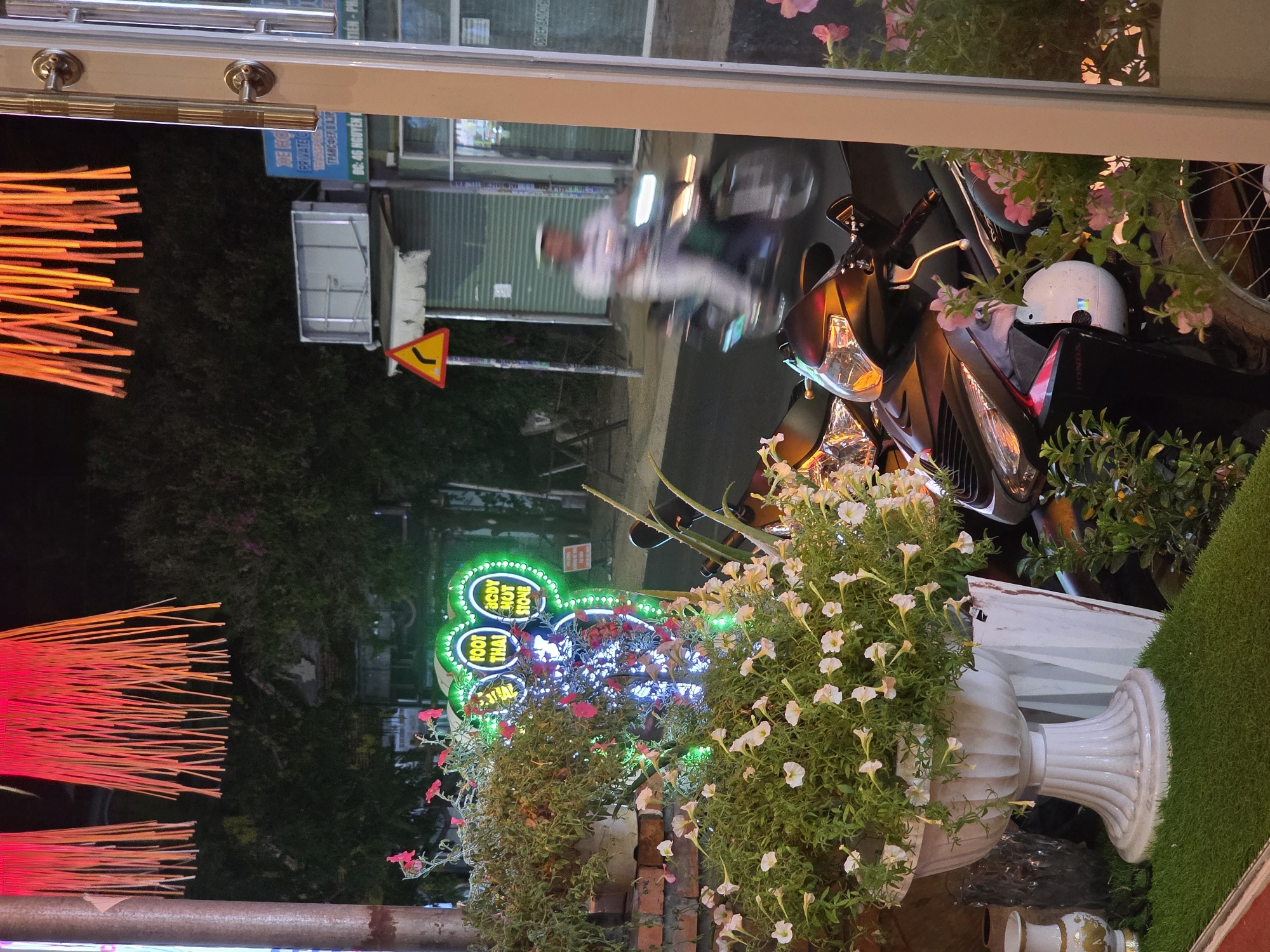 The scene is a night view, likely through a window, of a street in Phu Thuy Ward, Vietnam, featuring various elements. A motorcycle is parked on the right side of the frame, with a white helmet resting on its seat. Reflected in the window, a man is riding a motorcycle down the street. A sign partially reads "XE HO PRIVATE" and "ĐC: 46 NGUYỄN". A traffic sign indicating a curve in the road is visible. A green neon sign with the words "FOOT THAI" and "BODY HOT STONE" is prominent. An arrangement of flowers in a white, classical-style vase sits to the front. Orange and red light installations are situated along the left side of the frame. There is also artificial green grass next to the flowers in the vase.
