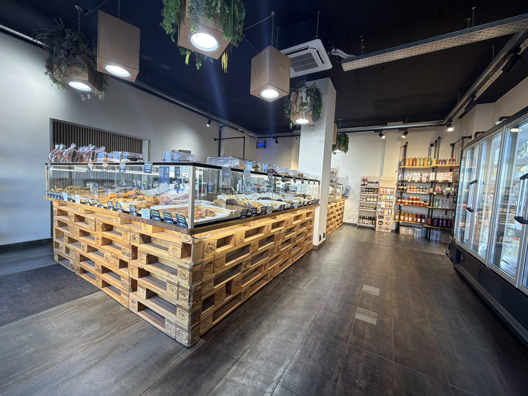 Here's a detailed description of the image:
**Location and Environment:** The image appears to be taken inside a bakery or deli, possibly in Sofia, Bulgaria (based on the presence of Cyrillic script on product labels). The interior features a dark ceiling with hanging light fixtures encased in brown paper-like boxes, some with greenery accents. The floor is tiled in a dark gray/brown tone.
**Objects:**
* **Display Counter:** A large display counter dominates the foreground. Its base is constructed from stacked wooden pallets with "EPAL" stamped on the sides. The counter itself has glass display cases containing various baked goods and pastries.
* **Shelving:** Along the right wall, there are shelves stocked with jars, bottles, and other packaged food items.
* **Refrigerated Display Cases:** Further to the right, there are large refrigerated display cases.
* **Entrance:** A doorway or entrance is visible in the background.
**Subjects:**
* There are no people visible in the image.
**Actions/Interactions:**
* The scene depicts a commercial space ready for customers but currently unoccupied.
**Overall Impression:** The image showcases a modern, slightly rustic interior of a bakery or food store with a focus on displaying baked goods and other products. The lighting, pallet-based counter, and shelving create a specific ambiance.