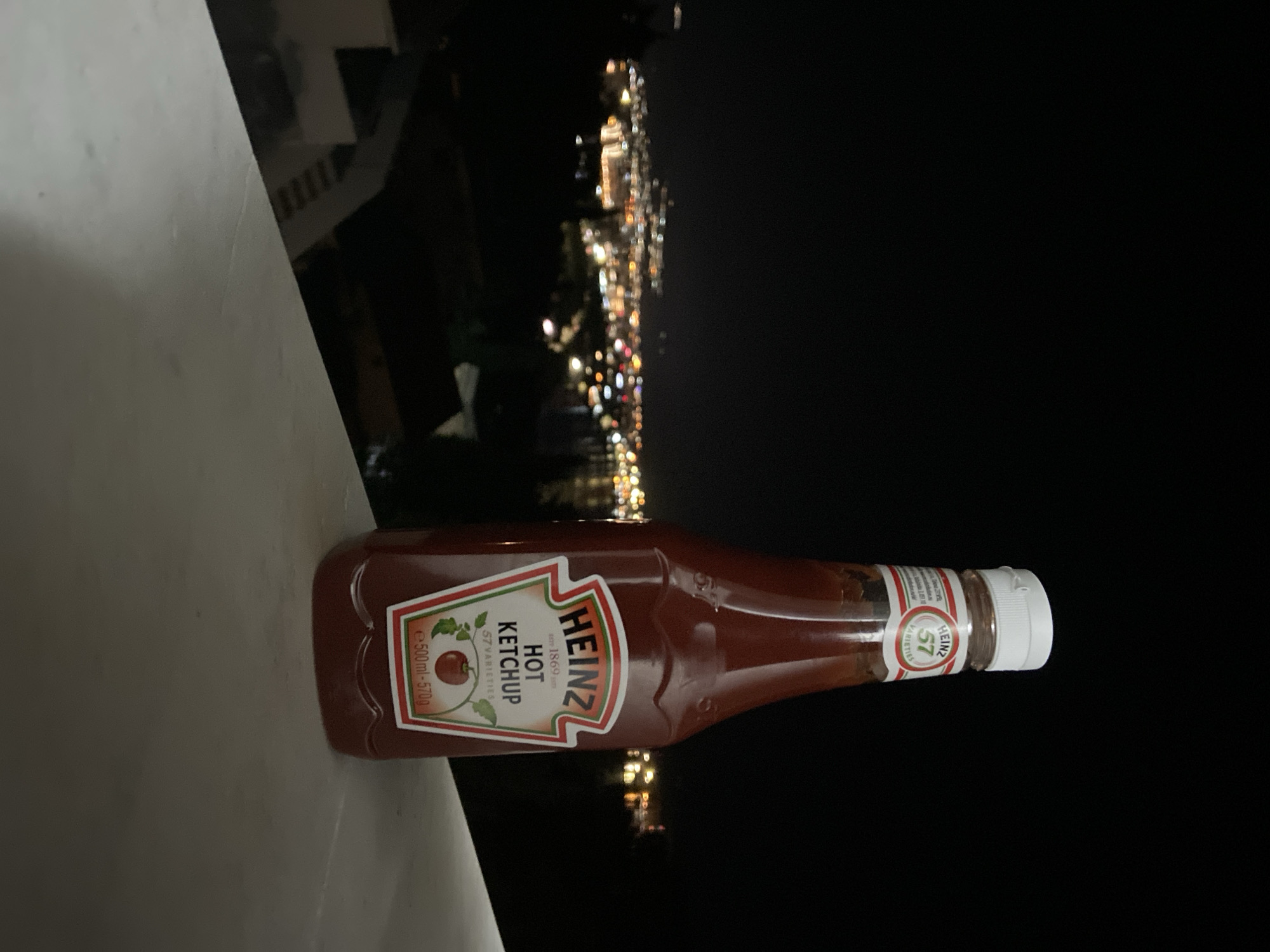 A bottle of Heinz hot ketchup lies on a white surface, likely a balcony railing. The background shows a dark night sky with blurred lights suggesting a distant cityscape. The blurry lights suggest the city of Šušanj, Montenegro, viewed from an elevated position at night. The bottle is lying horizontally, with the label facing the viewer, showing the Heinz logo, "Hot Ketchup," and a tomato image. The bottle has a white cap.