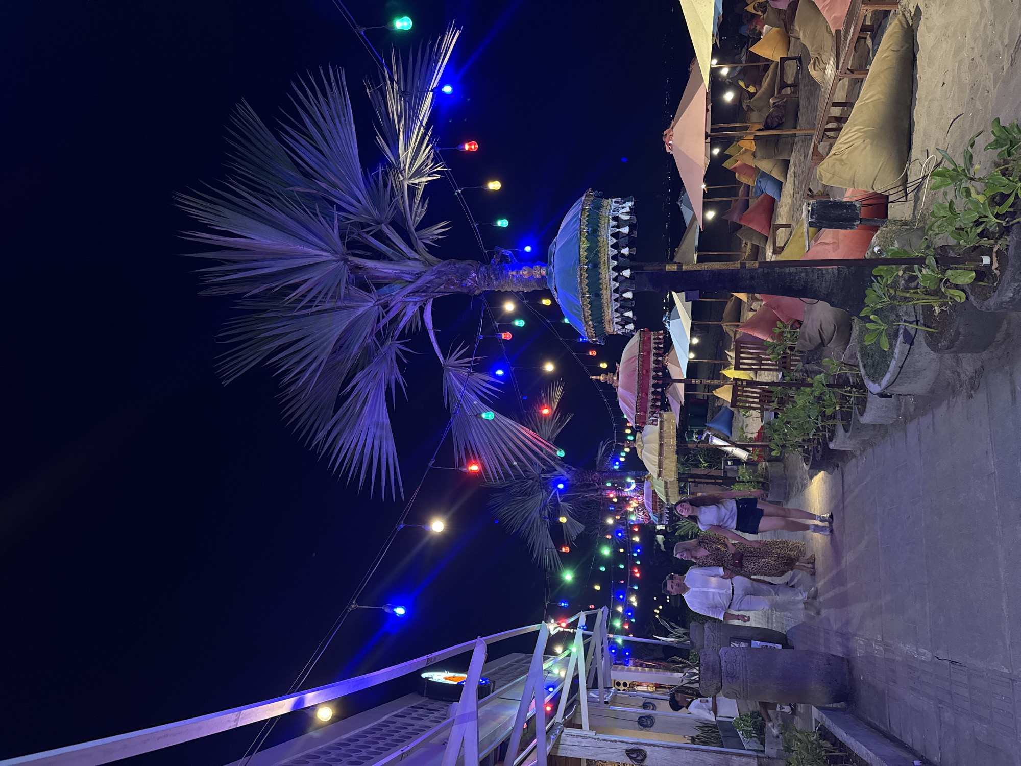 The image depicts a beachfront scene at night in Kuta, Indonesia, likely a popular dining or relaxation area. A walkway paved with grey bricks runs along the left side of the frame, bordered by potted plants and large stone planters. A building with white walls and a white slatted roof extends along the left edge, illuminated by strings of colorful lights (blue, green, red, yellow) that are draped across the scene.
On the walkway, three people are walking towards the viewer: a man in a white long-sleeved shirt and light-colored pants, a woman in a patterned dress, and a woman in a grey t-shirt and a dark skirt. To the right of the walkway, the scene opens up to a sandy beach. Numerous colorful beanbag chairs are scattered across the sand, arranged around low wooden tables and beneath beach umbrellas. A tall palm tree with fronds illuminated by blue light stands prominently in the mid-ground. Further back, more beach umbrellas and beanbags are visible, and in the distance, a faint outline of buildings or lights can be seen against the dark sky, suggesting the presence of the town. The overall atmosphere is vibrant and festive, with the colorful lights creating a warm glow on the beachside setup.