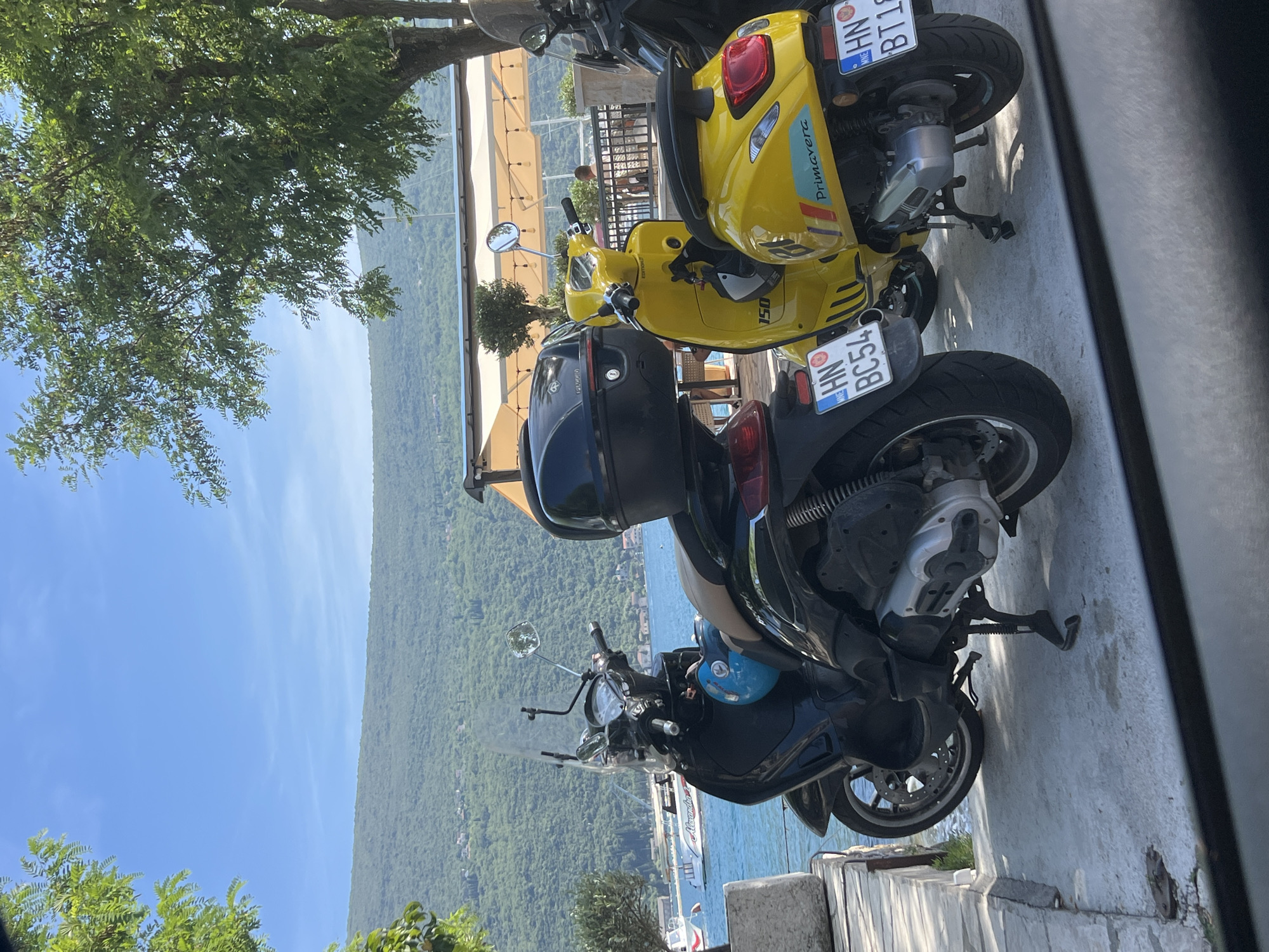 A bright, sunny outdoor scene captured in Đenovići, Montenegro, depicts a row of parked scooters by a waterfront promenade, with lush green mountains and clear blue skies in the background. The image is oriented vertically, showing the horizon line running down the middle.
In the foreground, several scooters are parked on a paved surface. Closest to the viewer is a black scooter with a top case, and a blue helmet visible near its handlebars. Further back are two yellow scooters. One yellow scooter clearly displays the text "Primavera" and "150" on its side, identifying it as a Vespa Primavera model. All scooters feature Montenegrin license plates. The black scooter has "HN BC54", and the yellow scooter behind it shows "HN BT18". "HN" indicates the registration city of Herceg Novi, which is in the Bay of Kotor area, near Đenovići.
Behind the scooters, a calm body of water, likely the Bay of Kotor, stretches across the midground. A white boat with "Aeronauta" visible on its side is docked or sailing on the water. On the opposite side of the bay, verdant, tree-covered mountains rise dramatically under a clear blue sky with a few wispy clouds.
To the right of the scooters, part of an outdoor seating area or cafe is visible with light-colored awnings and string lights. A person can be seen in the background near these structures. Large, leafy green trees frame the top left and bottom left corners of the scene, casting dappled shadows, suggesting a warm and pleasant day. The overall impression is one of a tranquil coastal town on a beautiful day.