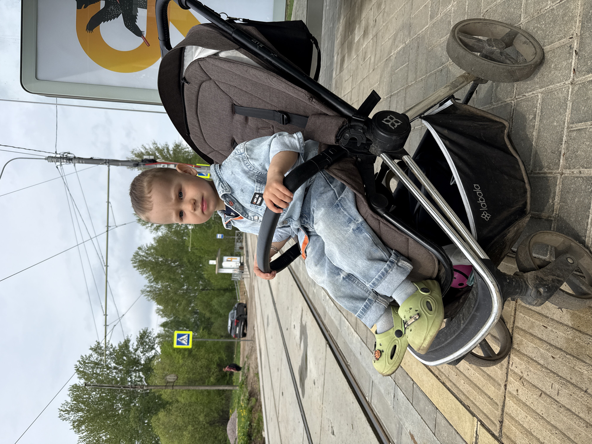 A young child sits in a black and brown stroller on a sidewalk, presumably in the city of Rusanovo, Russia, given the prompt's request. The child is wearing a denim jacket and light green Crocs. The sidewalk is partially composed of concrete slabs with parallel grooves running along their length, suggesting proximity to a roadway or traffic area. In the background, there's a yellow pedestrian crossing sign, trees, and power lines. A sign with a black raven on a yellow background is also partially visible. The perspective of the photograph gives a sense of looking down at the stroller from a slightly elevated position.