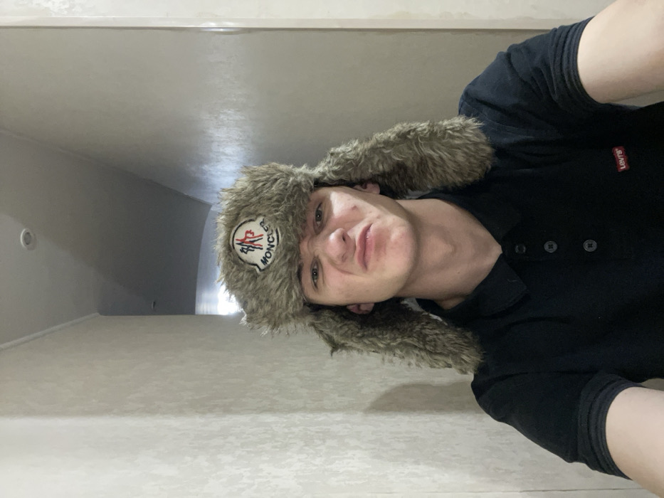 A man wearing a fur hat with a Moncler logo poses in an interior space. He's wearing a black polo shirt with a Levi's tag visible. The room is dimly lit, with a light-colored ceiling and wall. The perspective is skewed, giving a slightly disorienting feel. His expression is somewhat neutral or quizzical. The image is taken from a low angle, looking upwards towards the man.