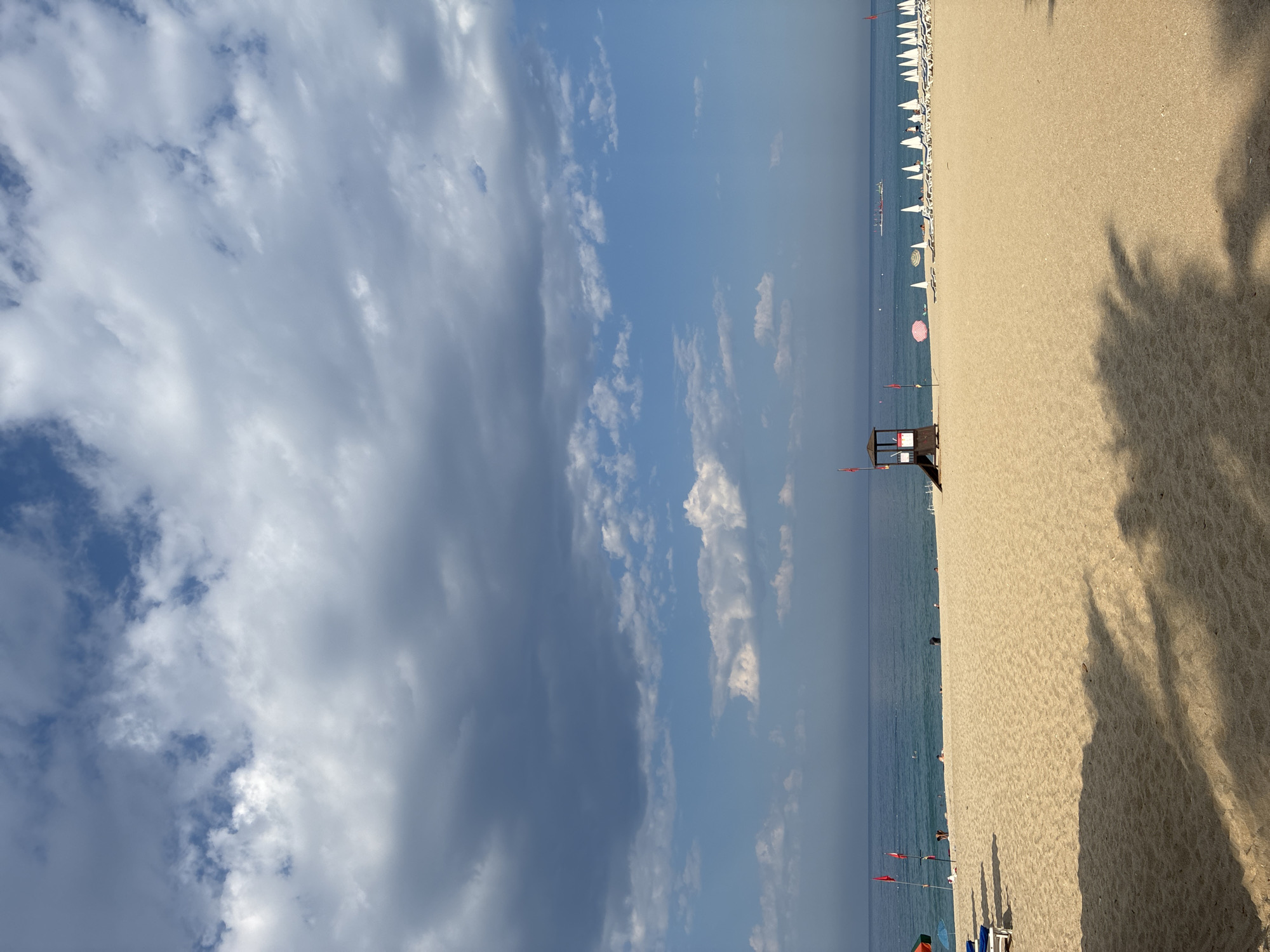 The image depicts a wide, sandy beach with the blue sea and sky in the background. A lifeguard tower stands on the left side of the beach, near a few red flags and umbrellas. In the distance, a row of white umbrellas and loungers are set up on the sand, indicating a populated beach area. There are a few people scattered across the beach and in the water. The sky is filled with a mix of dramatic, dark clouds and patches of bright blue sky with scattered white clouds. The overall scene is characteristic of a sunny day at a beach resort, likely in Alanya, Turkey, given the context.