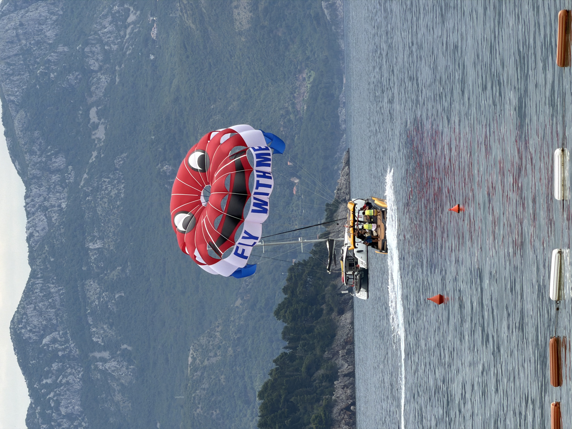 The image captures a parasailing scene off the coast near Budva, Montenegro. A parasail with a distinctive red, white, and blue design, emblazoned with the words "FLY WITH ME," is being towed by a yellow and white motorboat. The boat has several visible passengers on board. The water shows visible trails from the boat's wake. In the background, a mountain range covered in greenery rises steeply from the coastline. In the foreground of the water, there are orange buoys, with white and brown cylindrical floats attached, presumably marking a designated zone.