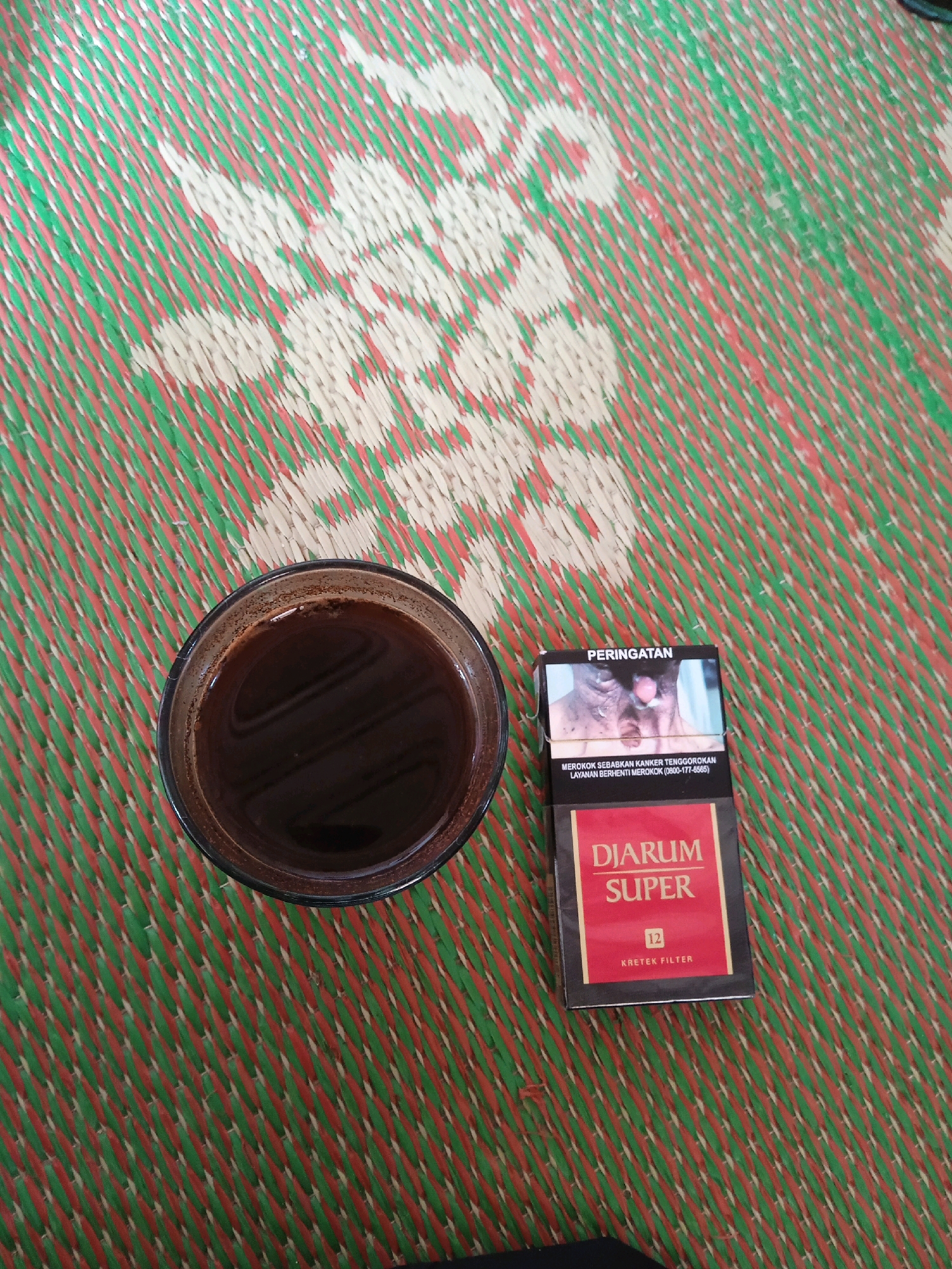 An overhead, medium shot captures a glass of dark coffee placed to the left of a pack of Djarum Super cigarettes on a woven mat. The mat has a diagonal weave pattern of red, green, and white strands. The coffee in the clear glass is dark brown and fills the glass nearly to the brim, with a thin layer of foam on the surface. The cigarette pack is oriented vertically with the brand name "DJARUM SUPER" prominently displayed in white lettering on a red background. Above the brand name, there is a warning image and text in Indonesian, including "PERINGATAN" (Warning). The number "12" is also visible on the pack, along with "KRETEK FILTER." The overall scene suggests a moment of relaxation or a break. The location is specified as Kelurahan, Indonesia.