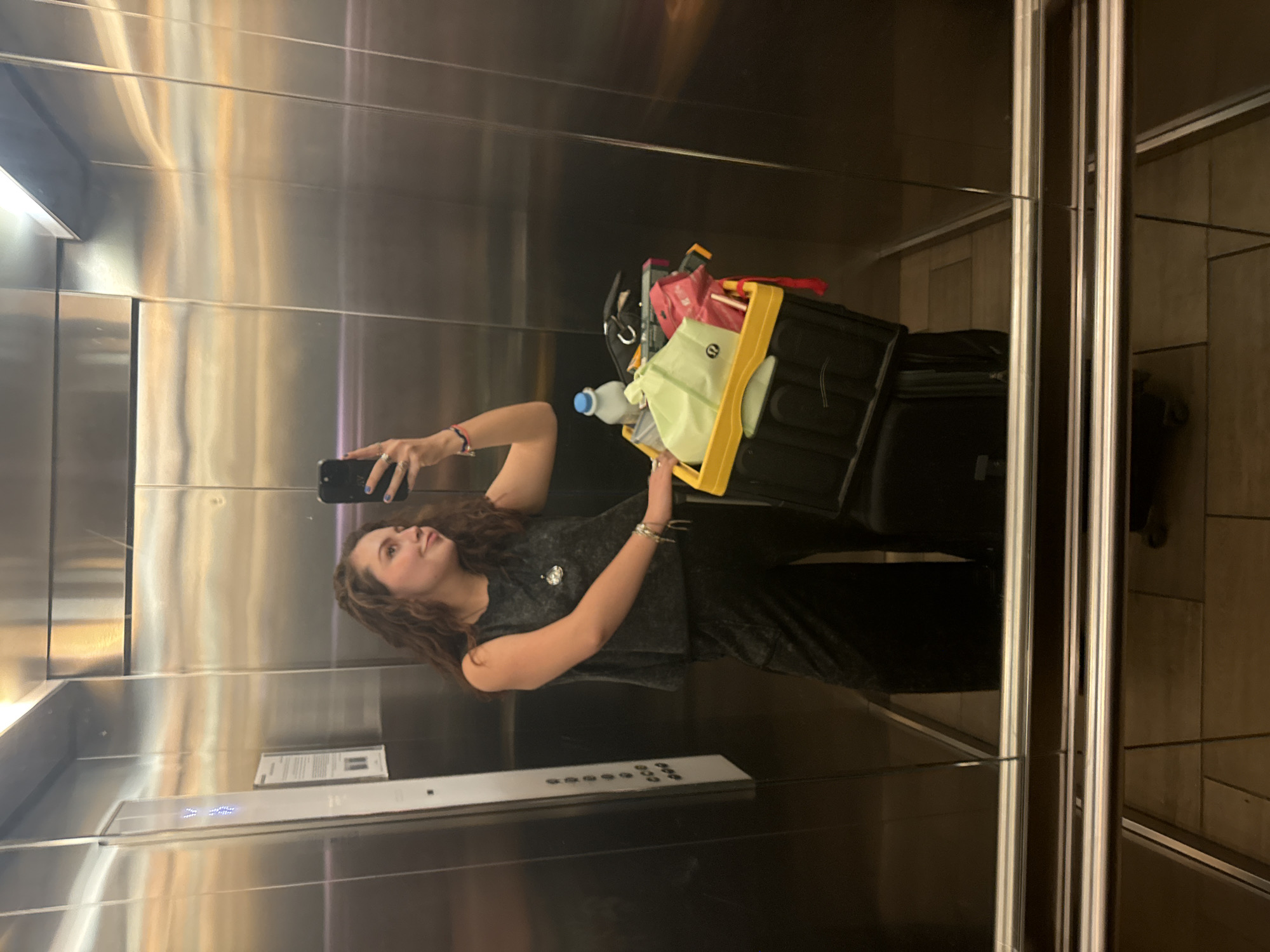 A woman is standing in an elevator taking a picture of herself in the mirror. She has long, curly brown hair and is wearing a gray tank top and dark pants. She is holding a black cell phone in her right hand. To her right is a black suitcase with a cart, which has various items, including a water bottle, a green and white bag, and a yellow container. The elevator has a mirrored interior with stainless steel paneling and controls. The location appears to be in Mexico City, Mexico.