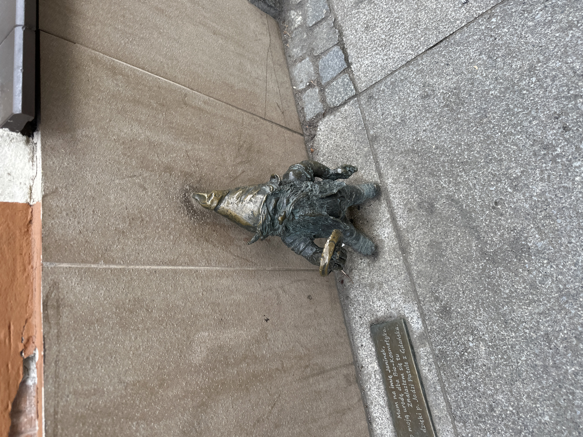 A ground-level, tilted, high-angle shot captures a small bronze statue of a gnome, typically found in Wrocław, Poland. The gnome lies on its stomach, head pointing to the left, near the junction of two types of pavement: one composed of large, smooth, light-brown tiles and the other, smaller grey stones. A short rectangular plaque with Polish text is positioned to the right of the gnome, slightly obscured by its hand. Close to the top-left corner, there is a building with a painted brown wall and a grey gutter. The statue appears somewhat aged and weathered. The text on the plaque translates to "My name is Janinek, Bio-Kosmetyka cares for my beauty. I found myself here thanks to Mrs. Jadzia Patale from Gdańsk."