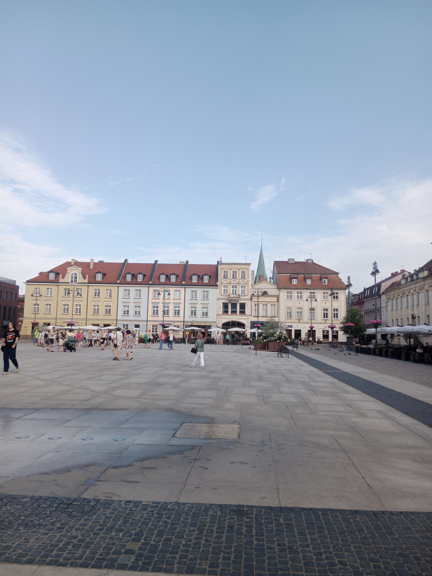 The image shows a wide, paved plaza in front of a row of colorful European-style buildings. The buildings are multi-story with red-tiled roofs and many windows. Some of the buildings have awnings or umbrellas set up in front, suggesting outdoor seating for cafes or restaurants. A tall, green spire rises from one of the buildings in the background. The plaza is sparsely populated with several people walking and standing around. The ground is paved with large stone tiles and in the foreground, there are cobblestones. The sky is a pale blue with some scattered white clouds. The overall impression is of a pleasant, sunny day in a historic European city square.