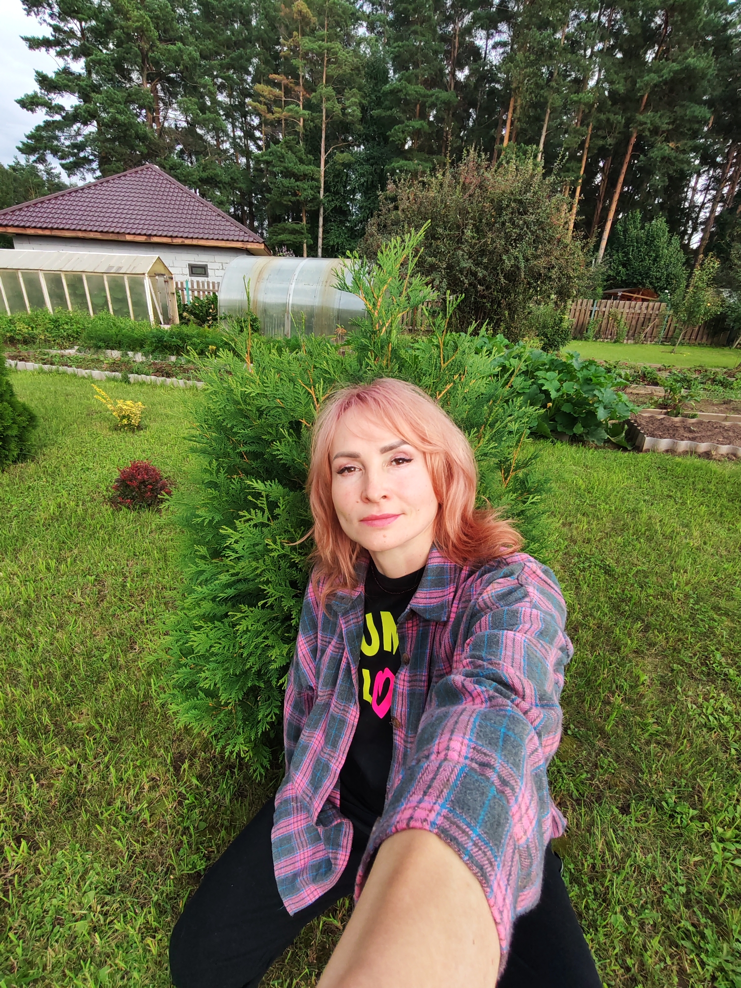 A young woman with light pink hair and wearing a pink and gray plaid shirt over a black t-shirt is sitting in a grassy yard. She is holding her phone out to take a selfie. Behind her, a large green bush is visible. In the background, there is a greenhouse, a garden with raised beds, and a wooden fence. Tall pine trees are also visible behind the garden. The scene appears to be a suburban or rural area, possibly a dacha in Chaikovsky, Russia.
