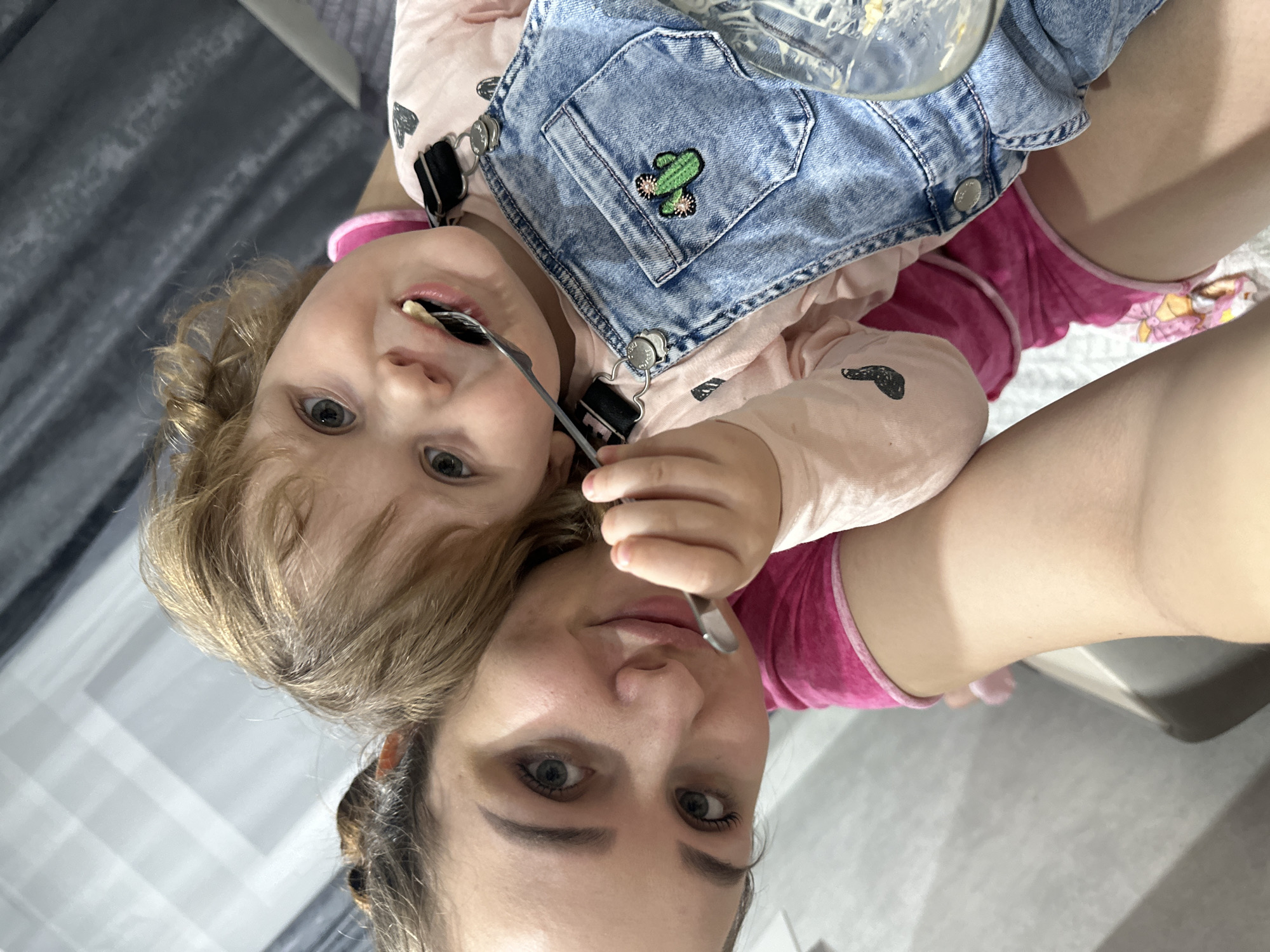 A woman is holding a young child. The child is wearing denim overalls with a cactus patch on the pocket and a long-sleeved pink shirt. The child is eating something from a spoon. The woman is holding the child and looking at the camera. The location appears to be indoors. There is not enough information to determine the city or any other location in Russia.