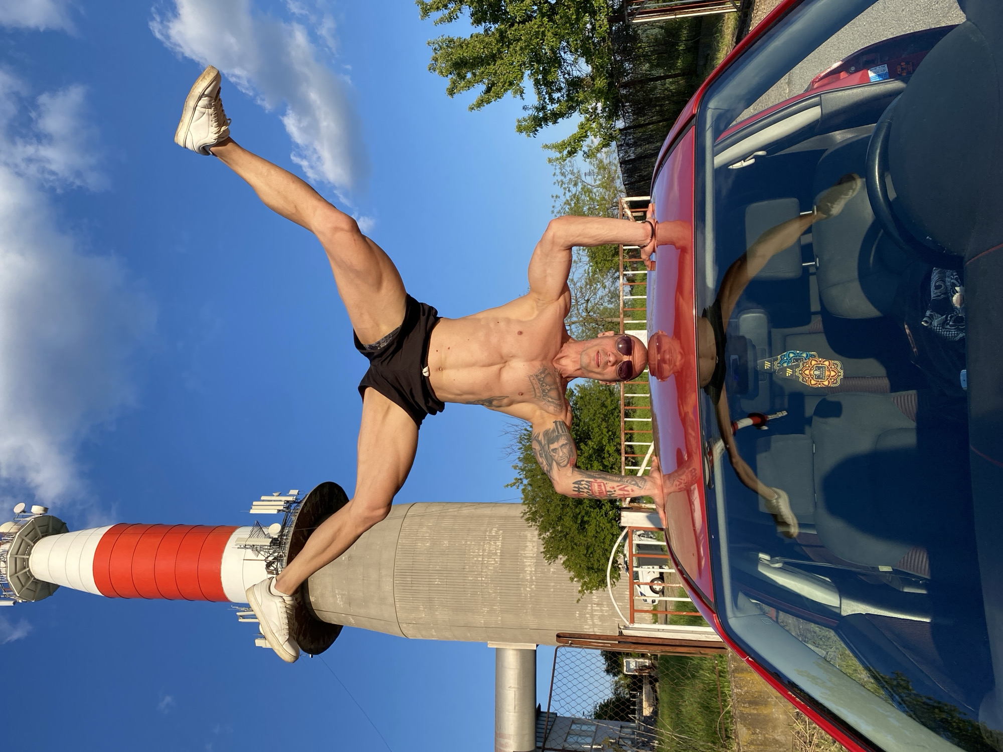 A man in black shorts and white sneakers is performing a handstand on a red car. His left leg is extended towards a tall, cylindrical tower with alternating red and white stripes, topped with various antennas and communication equipment. The backdrop includes a blue sky with scattered clouds, trees, and parts of a fence and building. The car's interior, including seats and a steering wheel, is visible through the windshield. The location is Pleven, Bulgaria, identifiable by the tower's design and function as a telecommunications landmark.