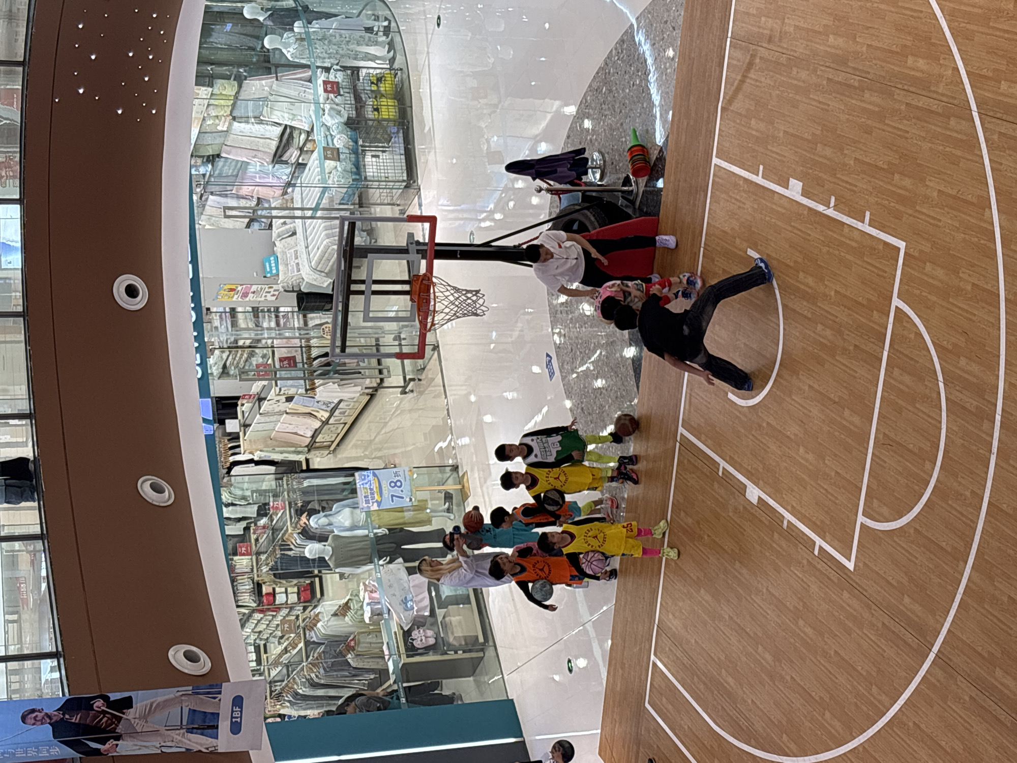 The image depicts an indoor basketball court within a shopping mall in Jiangjin, China. A group of children in sports attire are participating in a basketball activity. Adults are also present, seemingly instructing or supervising the children. A basketball hoop is visible. The background shows a retail area with mannequins and merchandise displays.