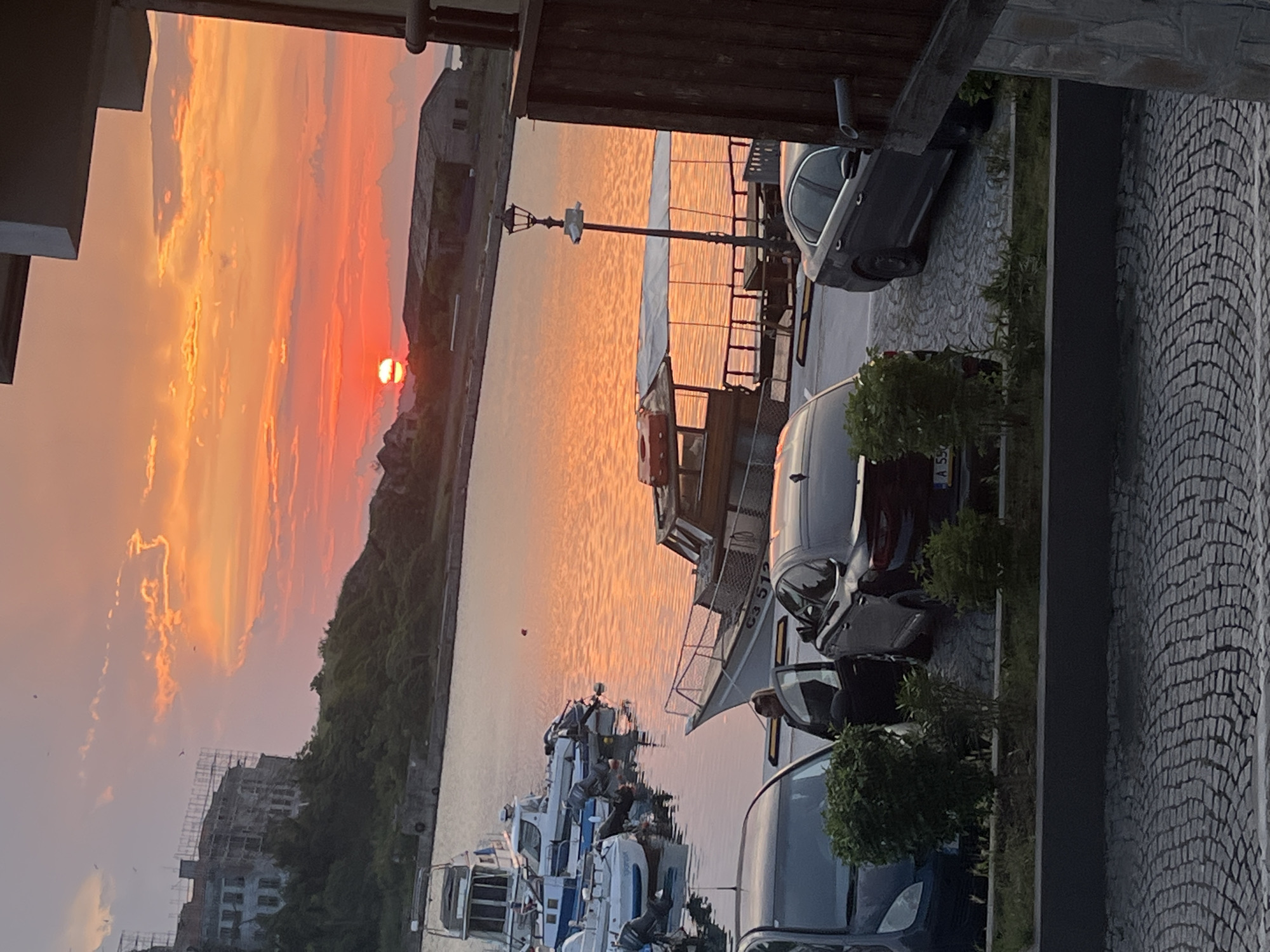 This media file depicts a vibrant sunset over a waterfront scene, likely a harbor or marina. The image is rich with warm hues of orange, red, and pink dominating the sky and reflecting intensely on the calm water.
The sun, a distinct fiery red-orange orb, is positioned low on the horizon on the left side of the frame, casting long, dramatic light across the clouds. The water, possibly a sea, lake, or river, reflects these brilliant colors, creating a glowing path extending towards the foreground.
Numerous boats are docked in the water. In the mid-ground, a larger, darker boat with a cabin is prominent, displaying "C3 512" on its side, while several smaller, white recreational boats or speedboats are also visible. A street lamp post stands near the water, and a small white object, possibly a bird, is perched atop it.
On the distant left horizon, a large building under construction with scaffolding is visible, along with some trees. Further right on the distant shore, another older, dark-roofed building can be seen. The foreground on the right features a paved area with a distinctive cobblestone pattern, indicative of a walkway or road. Several cars are parked along this path, including a dark sedan with a partially visible license plate "A 55..." and other vehicles. Small green shrubs line the paved area.
No people are visible in the image. The overall atmosphere is tranquil and picturesque, capturing the peaceful end of a day at a waterfront location.