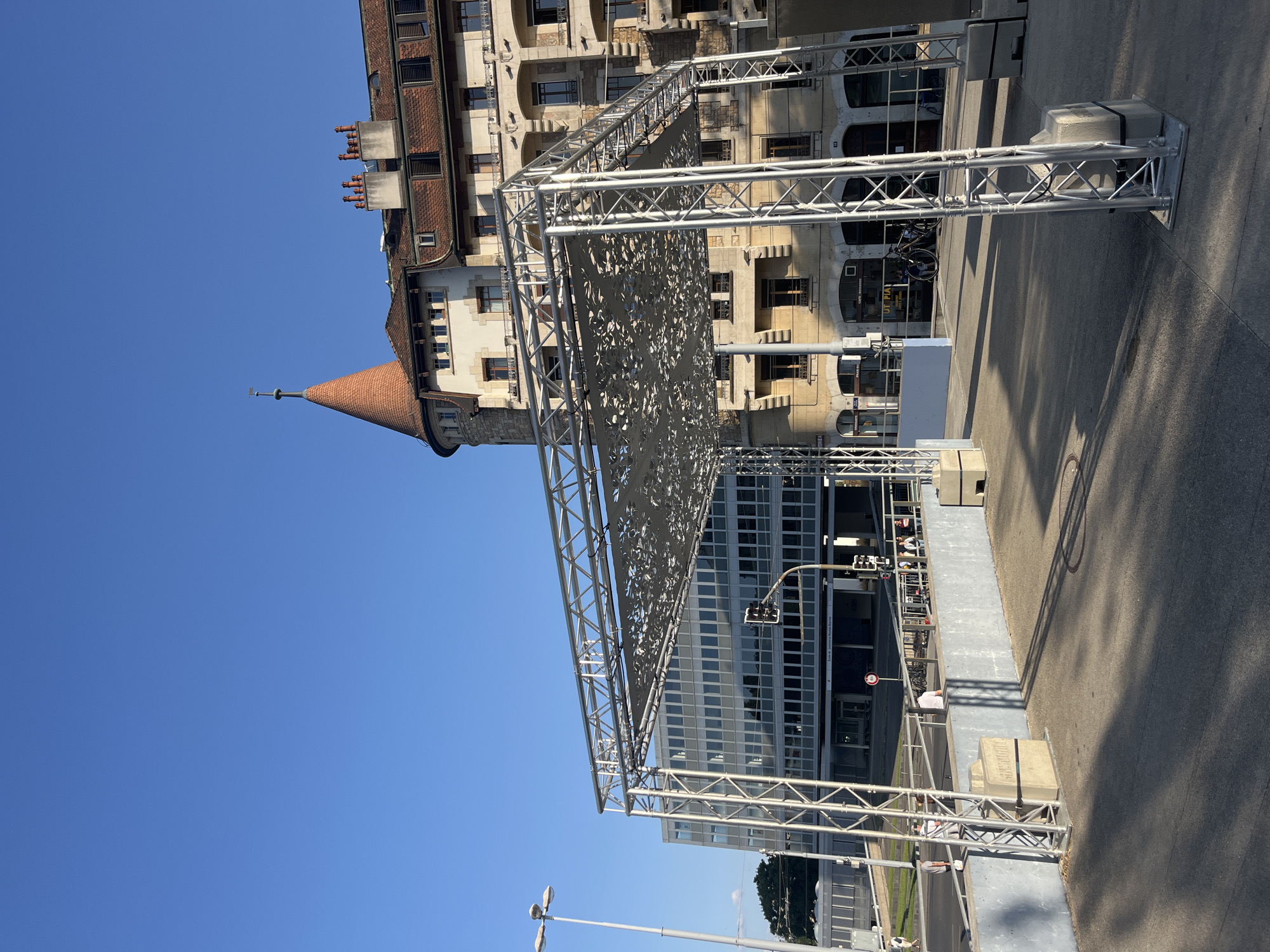 This is a photograph of an outdoor structure in Geneva, Switzerland. The structure is a large, metallic scaffolding with a decorative, patterned fabric canopy. It is situated in an urban environment, with a modern glass-fronted building and an older, multi-story building with a conical turret in the background. A street with a traffic light and several people are visible in the middle ground, and a clear blue sky fills the upper portion of the image. The lighting suggests it is daytime, likely late afternoon due to the shadows. The scaffolding appears to be temporary, possibly for an event or installation.