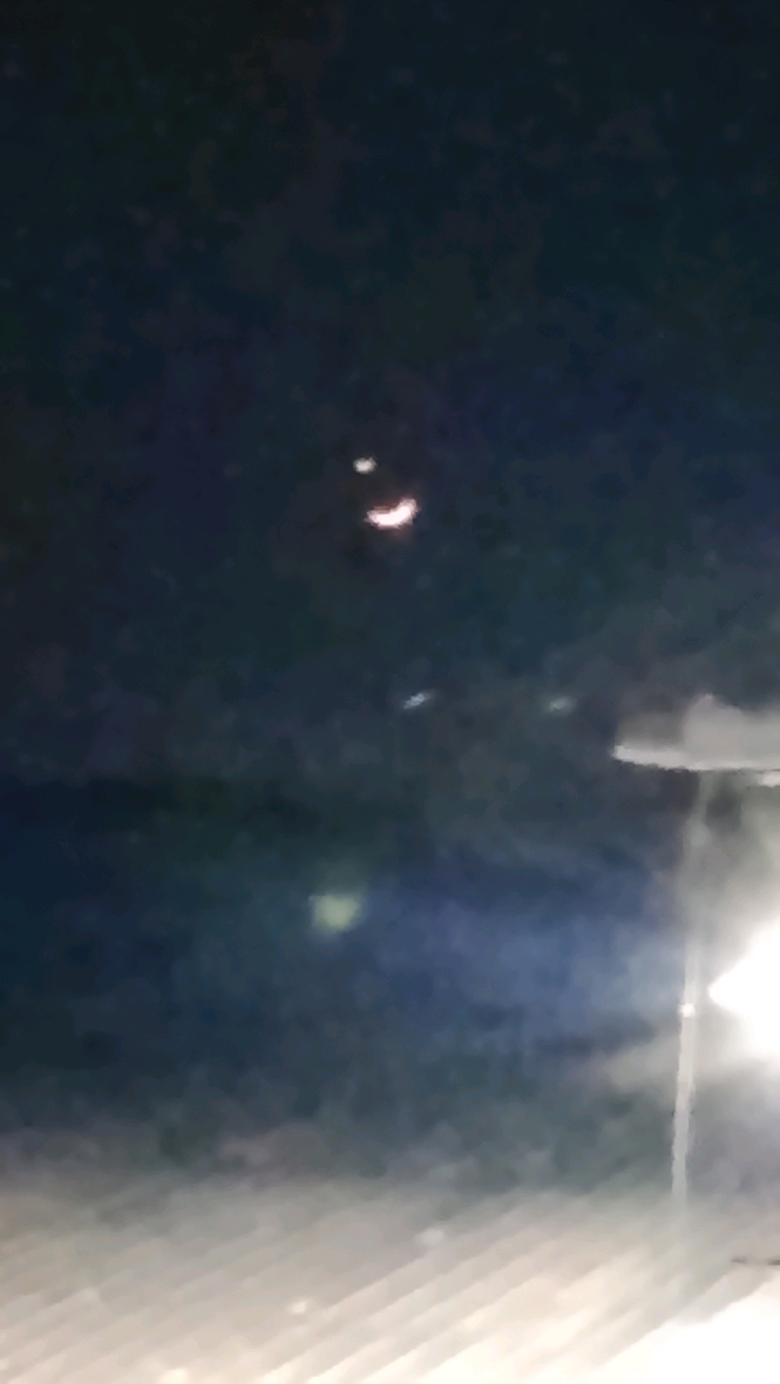 The image shows a dark, blurry night sky with several lights. In the upper center, there are two small, bright lights, one slightly above and to the left of the other, resembling a crescent moon shape. Below these, a faint, elongated white light is visible. Further down and to the left, a blurred yellowish light is present. The lower portion of the image is dominated by a blurry, lighter area that appears to be illuminated by artificial light sources, possibly a streetlamp or building lights, casting shadows and creating a hazy effect. The overall scene suggests an outdoor environment at night, possibly within or near a city due to the presence of artificial lights. There are no discernible people or specific landmarks to pinpoint the location within Ujungrusi, Indonesia, beyond it being an outdoor, nighttime setting.