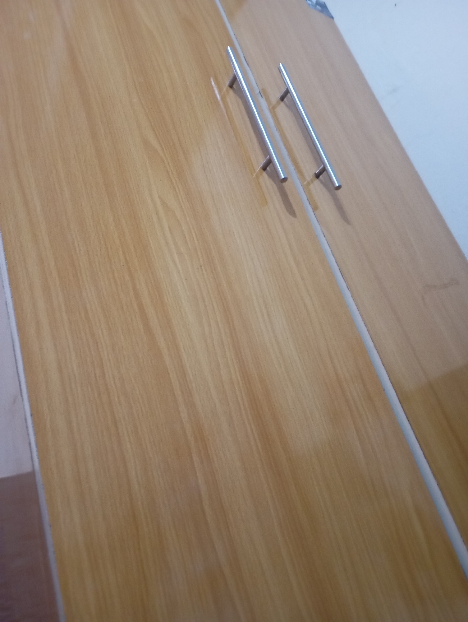 Two wooden closet doors with silver handles are visible in the image. The doors are oriented diagonally from top left to bottom right, with the left door taking up most of the frame. The wood grain is light brown with darker brown lines running vertically. The handles are long and cylindrical, each attached to the doors with two perpendicular supports. The right door is partially visible on the right side of the image. The background is a plain white wall.