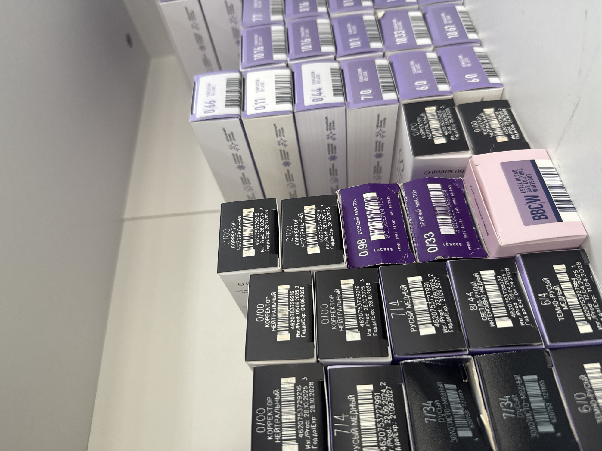 The image shows a variety of hair dye boxes, primarily of the brand "Estel", arranged on a white surface, possibly within a store or storage area.
The dyes are categorized by shade and purpose, with boxes labelled "Sensation de Luxe" and "Correction". Specific shades include "0.66", "0.11", "0.44", "70", "6.0", "10.16", "77", "816", "10.33" and "10.61". There are also corrector dyes labelled "0/00" (Neutral), color mixer dyes "0/98" (Pink) and "0/33" (Green). Other dyes include "7/14", "8/44", "6/4", and "7/34".
One box is specifically identified as "Estel Blond Bar Couture Whiteout" (BBCW), a hair lightening cream.
Information printed on the boxes indicates the dyes were produced and expire around 2024-2028. There are some Russian words indicating the products were manufactured for the Russian market.
There is no visible evidence of a location, city or region in the image. Therefore, the city of Chaikovsky cannot be confirmed.